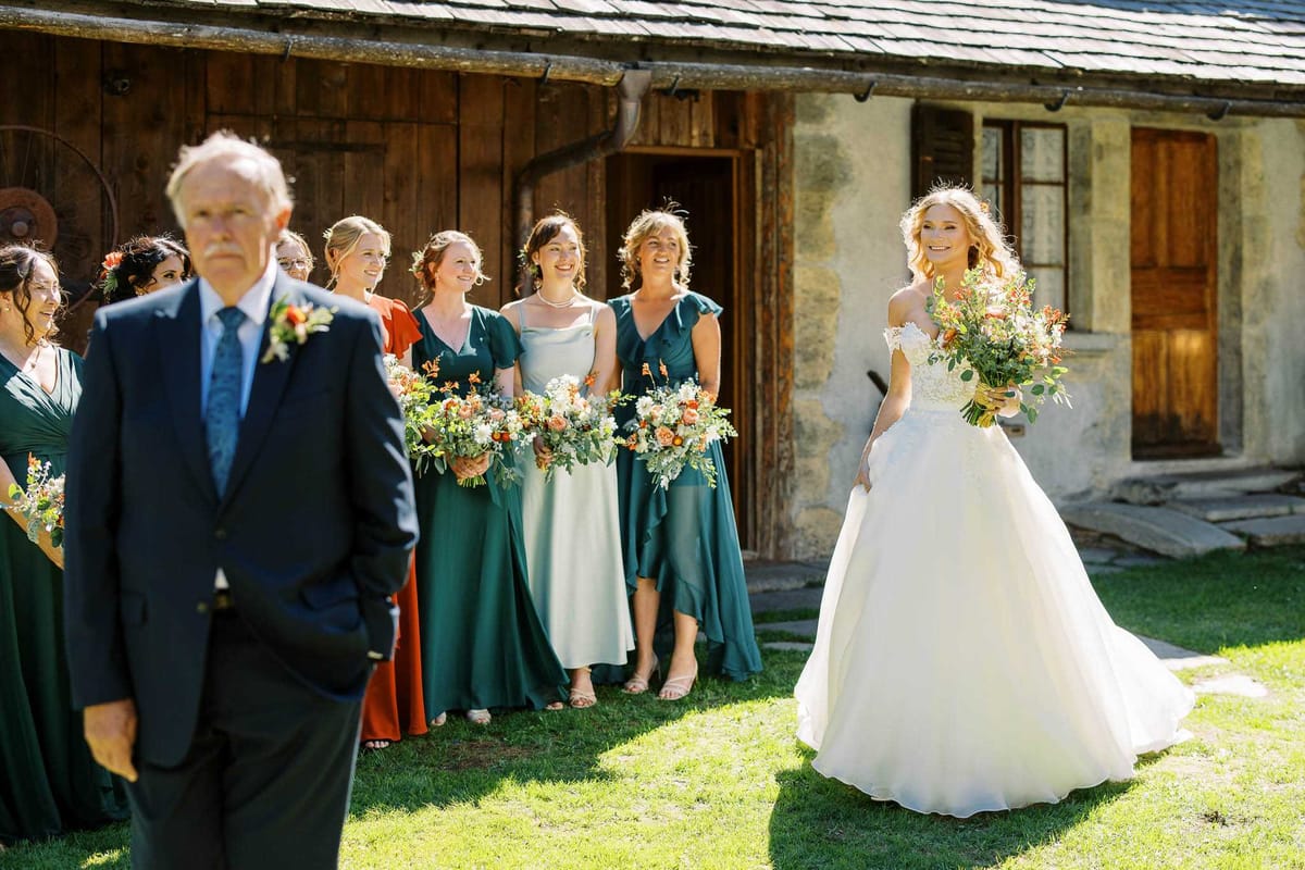 The bride and her bridal party are gathered outdoors in front of a rustic stone and timber building, appearing to prepare for the processional. The bride stands to the right wearing an off-the-shoulder ballgown with a full white skirt and lace bodice, holding a large bouquet of orange, coral, and white blooms with greenery. Approximately five bridesmaids are lined up behind her in mismatched green dresses ranging from deep forest green to sage and mint, each carrying coordinating bouquets of orange, coral, and cream flowers with eucalyptus. A man in a navy suit with a blue tie and an orange buttonhole — likely the father of the bride — stands in the foreground to the left with his back partially to the camera. The overall color palette is green and burnt orange with a relaxed, rustic styling, and the image is a wide candid shot taken in bright natural sunlight.