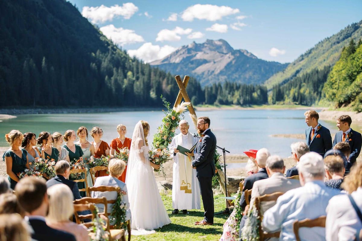 An outdoor wedding ceremony taking place on the grassy shore of a mountain lake, with the couple standing before an officiant dressed in white liturgical robes with a gold Alpha and Omega stole. The ceremony arch is a rustic wooden X-frame structure adorned with lush greenery, coral and terracotta blooms, and pampas grass. The bride wears a white gown with a long veil and holds a large bouquet of coral and orange flowers, while the groom wears a navy suit with an orange tie and boutonniere. The bridal party of approximately eight bridesmaids stands to the left, wearing a mix of teal/forest green and rust-orange wrap dresses and carrying matching floral bouquets; two or three groomsmen in navy suits stand to the right. Wooden folding chairs seat approximately 30-40 guests on either side of the aisle, with floral arrangements decorating the chair ends. The wide-angle shot captures the full ceremony scene with the turquoise lake and mountain peaks as a dramatic backdrop.