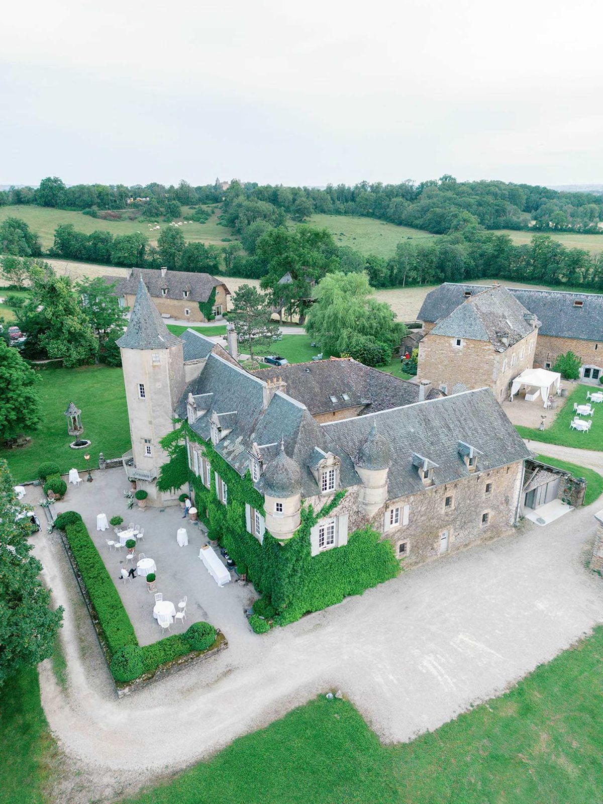An aerial drone shot of a French château wedding venue set within rolling countryside. The multi-wing stone château features slate grey rooftops, a round tower, and ivy-covered facades. In the courtyard to the left of the main building, white-linen-covered round tables and white chairs are arranged for an outdoor reception or cocktail hour, with a small number of people visible on the grounds. A white canopy tent is set up on the lawn to the right side of the property. The estate includes several outbuildings and is surrounded by manicured hedges, gravel pathways, and expansive lawns. Potential venue feature image.