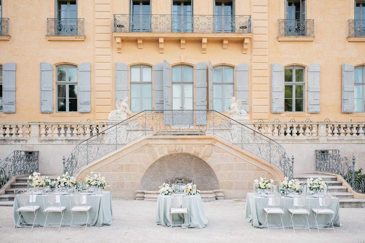 Outdoor reception setup in the courtyard of a French chateau, with three round tables dressed in sage green floor-length linen tablecloths arranged symmetrically in front of the venue's grand double staircase. The wrought-iron staircase features ornate scrollwork and is flanked by two white stone figurative sculptures, with the chateau facade displaying warm ochre-toned render, grey-blue shuttered arched windows, and iron-railed balconies above. Each table is styled with low floral centerpieces composed of ivory, white, and soft yellow blooms — likely roses, ranunculus, and similar garden flowers — alongside taper candles and glassware. The chairs are white metal with a medallion back design and white seat cushions, consistent with a classic French aesthetic. The wide shot captures the full architectural facade as the primary backdrop, with no people present. Potential venue feature image.