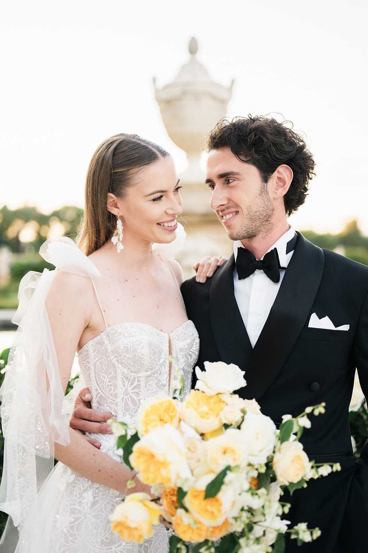 A couple portrait taken outdoors at golden hour, with a large stone urn architectural feature visible in the soft-focus background, suggesting a formal chateau or estate setting. The bride wears a fitted ivory lace corset-style gown with spaghetti straps and an oversized organza bow detail at the shoulder, paired with dangling pearl and crystal earrings and her hair pulled back sleekly. The groom wears a black tuxedo with a black bow tie and white pocket square, his arm around the bride's waist as both smile at each other. The bride holds a lush bouquet of golden yellow and ivory garden roses with small white stephanotis-style blooms and green foliage, providing a warm color accent against the white dress. Close-up portrait composition with a shallow depth of field.