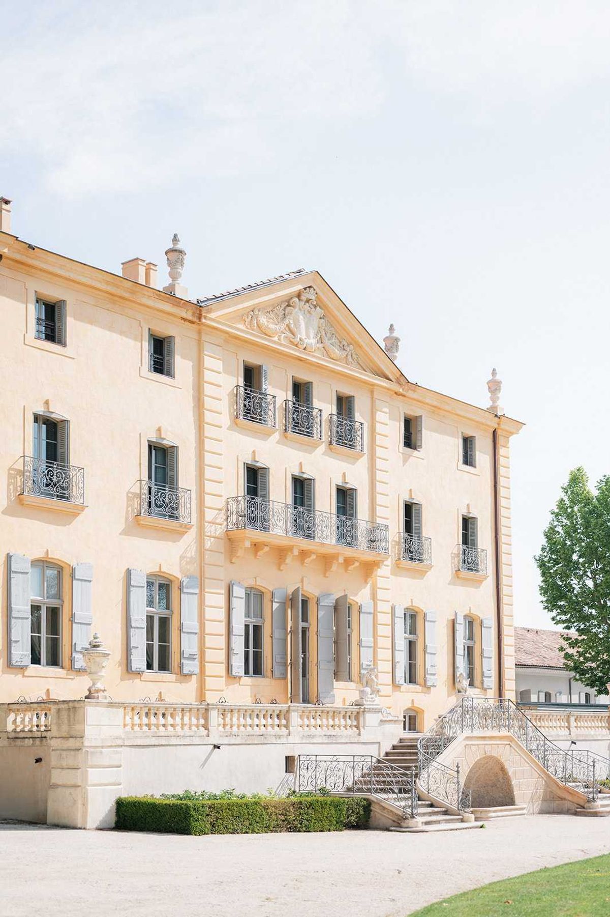Wide exterior shot of a large classical French or Italian-style manor house or château, featuring warm peach-toned stucco façade with grey-blue shuttered windows across three stories. The building displays ornate architectural details including a carved pediment with decorative relief at the roofline, wrought iron Juliet balconies, stone balustrade terrace, and a curved double staircase with wrought iron railings leading to the main entrance. Stone urns and sculpted figures accent the terrace, and trimmed box hedging lines the gravel forecourt. No people are visible; the image functions as a standalone architectural shot. Potential venue feature image.