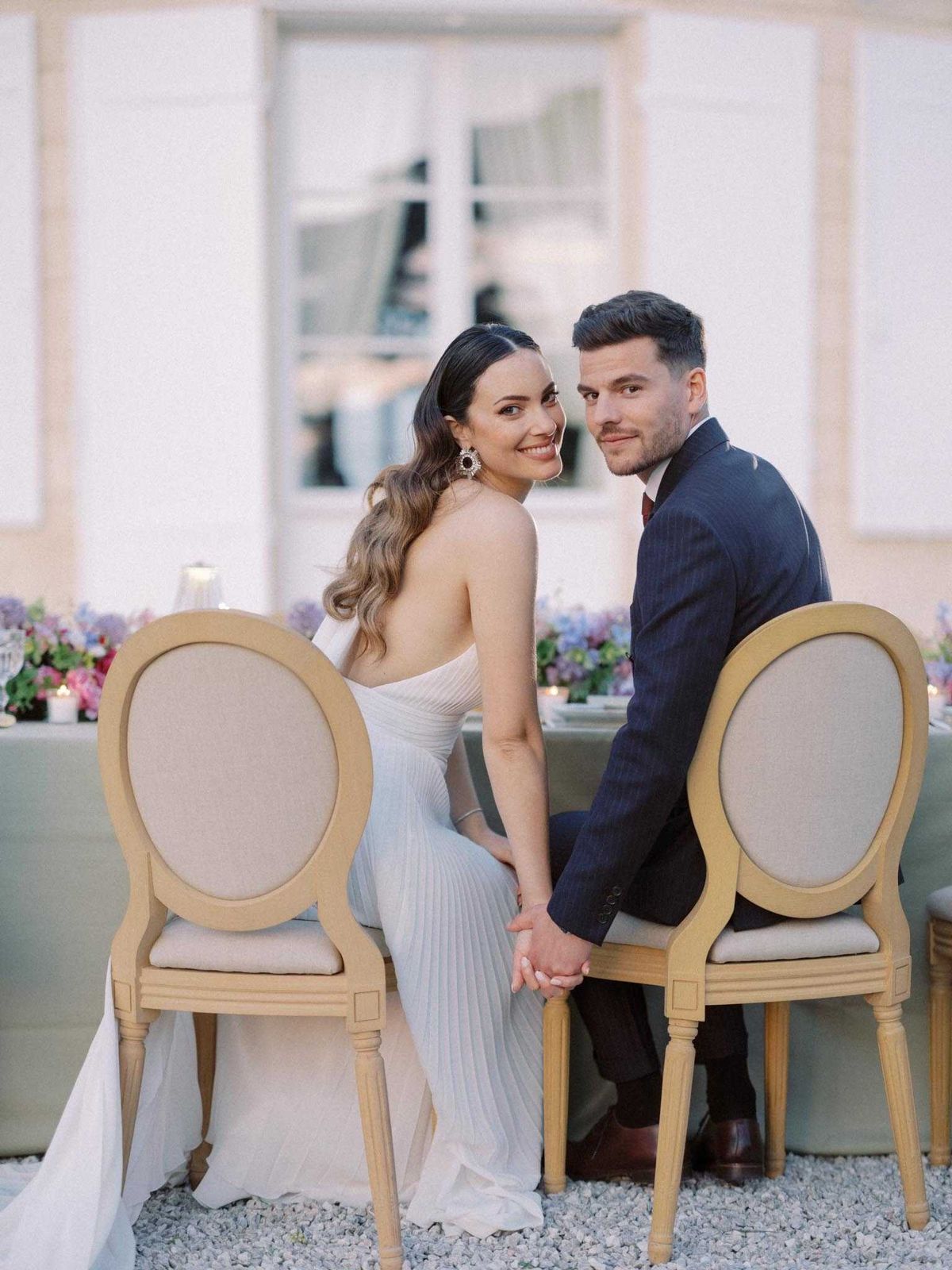 A couple portrait taken at an outdoor reception, with the bride and groom seated at their sweetheart or head table and turning back to face the camera while holding hands. The bride wears a strapless white pleated gown with a low back and long wavy hair styled to one side, accessorized with large statement earrings. The groom wears a navy pinstripe suit with a burgundy tie and brown dress shoes. They are seated on natural wood Louis XVI-style chairs with beige upholstered oval backs, positioned on a white gravel surface. Behind them, the reception table is dressed in a sage green linen with a lush floral runner featuring purple hydrangeas, pink and red blooms, and scattered pillar candles. The background shows the white stone facade of a French chateau with tall shuttered windows. The image is a medium portrait shot with shallow depth of field, keeping the couple sharp against the softly blurred venue and tablescape.