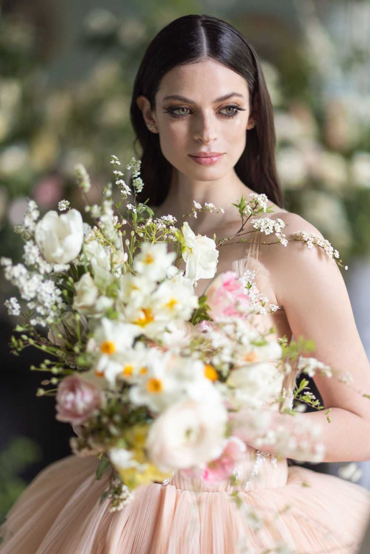 A close-up bridal portrait of a dark-haired woman wearing a blush peach pleated tulle gown with delicate floral or embellished shoulder straps. She holds a loosely arranged garden-style bouquet composed of white tulips, white daffodils with yellow centers, pink garden roses, blush ranunculus, and white blossom branches with fine green foliage. Her makeup features defined cat-eye liner and a neutral lip. The background is softly blurred with hints of white and pink florals, suggesting a floral-filled indoor or greenhouse-style setting. The composition is a shallow depth-of-field portrait with the bouquet partially out of focus in the foreground, drawing attention to the subject's face.