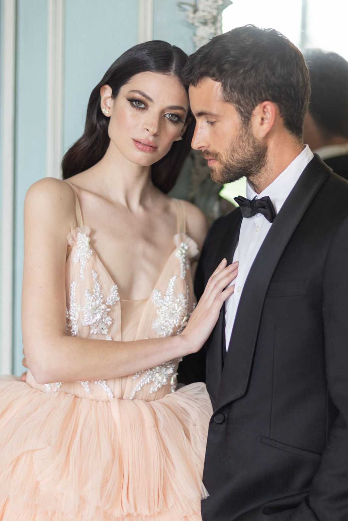 A close-up portrait of a couple posed together indoors against a pale blue-green wall. The bride wears a blush peach tulle ball gown with spaghetti straps and a deep V-neckline, featuring intricate white beaded and embroidered floral appliqué on the bodice; her dark hair is styled in loose waves to one side and she wears small crystal ear studs. The groom is dressed in a black tuxedo with a shawl lapel, white dress shirt, and black bow tie. The bride rests her hand on the groom's chest while leaning her head toward his, as he looks downward; the overall styling aesthetic is classic and formal. The shot is a tightly framed couple portrait with a shallow depth of field that keeps the background soft.