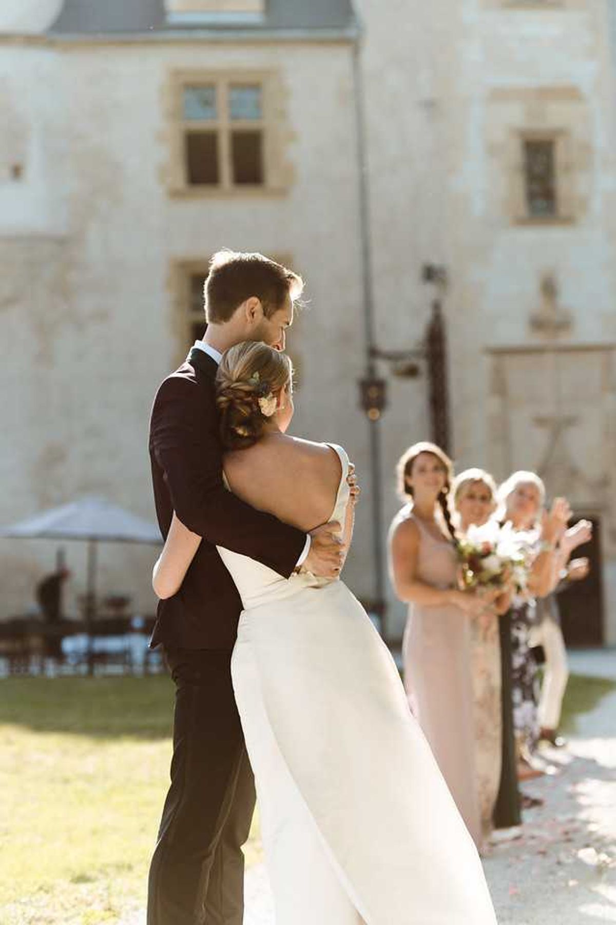 The bride and groom embrace outdoors in front of a French chateau, shot from behind in a medium portrait composition. The bride wears a low-back ivory gown with a fitted silhouette and has her hair styled in a braided updo with a small floral hair accessory, while the groom wears a dark navy suit. In the soft background, three women — appearing to be bridesmaids or guests — look on and applaud; one wears a blush pink dress and holds a bouquet with white and deep red blooms. The chateau facade with arched ironwork lanterns is visible behind the group, along with outdoor ceremony seating and a parasol to the left. The image is bathed in warm late-afternoon sun, giving it high contrast and a golden tone.