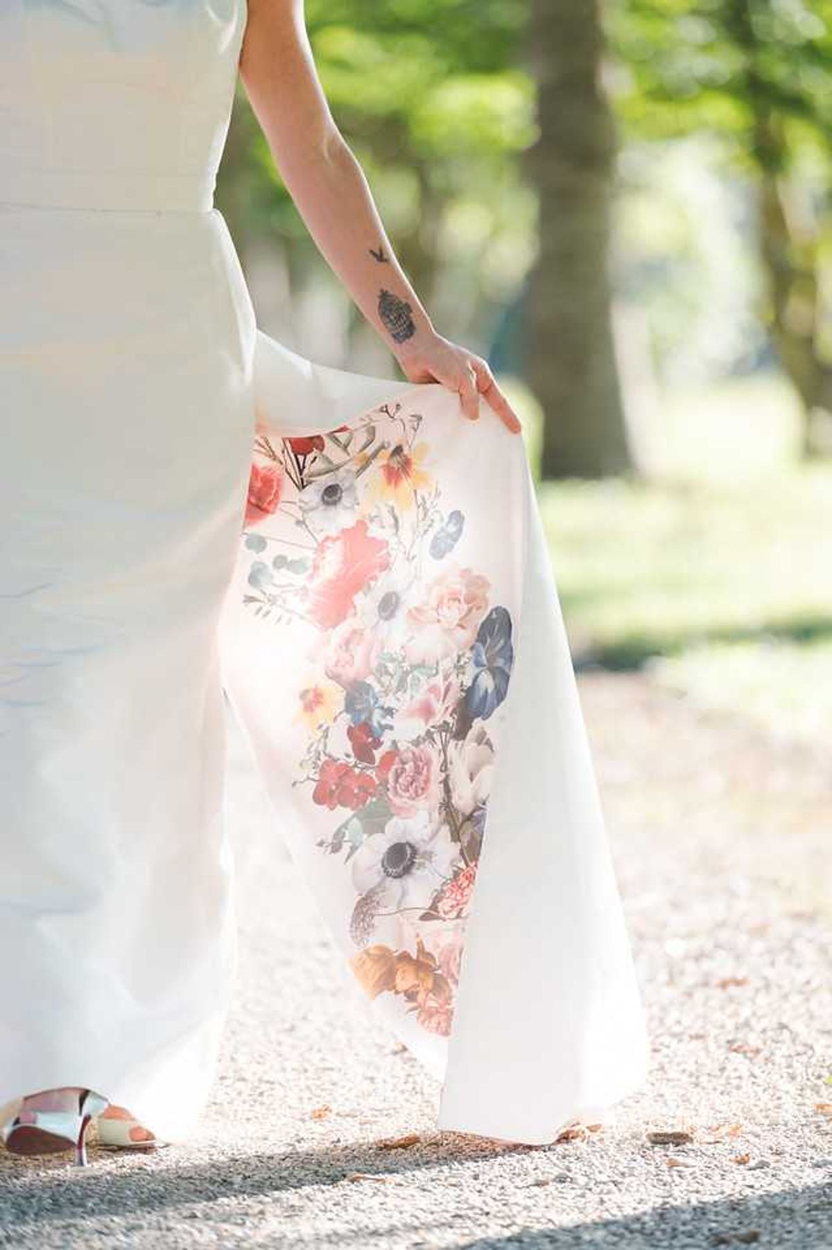 Close-up detail shot of a bride lifting the skirt of her white wedding dress to reveal a large floral print on the interior lining, featuring painted-style blooms in coral, blush pink, dusty blue, red, and yellow — including roses, anemones, and wildflowers. The bride has visible tattoos on her forearm and wrist, and is wearing metallic silver open-toe heels. The shot is taken outdoors on a gravel path in natural sunlight, with a soft bokeh background of trees.
