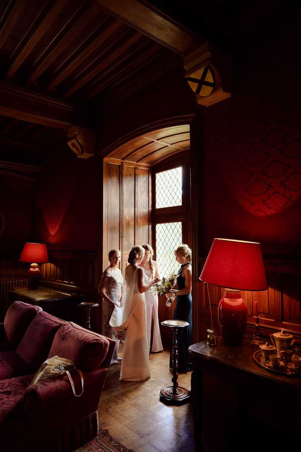 A getting-ready or pre-ceremony moment captured indoors in a richly decorated room of what appears to be a historic chateau or manor house, featuring dark wood-paneled walls, a carved arched window with leaded diamond-pane glass, exposed beam ceiling, parquet flooring, and deep red lampshades on ornate table lamps. Three women stand near the window in natural backlight: the bride wears a sleek ivory column gown and holds a white bouquet with greenery, flanked by a woman in a lace floral dress and a woman in a dark navy or black dress. The room is furnished with burgundy velvet armchairs, a sideboard with tea service, and decorated with damask-patterned red walls, giving the space a classic, richly toned interior. The wide-angle shot uses the dramatic contrast between the bright window light and the deep warm tones of the room's interior lighting.
