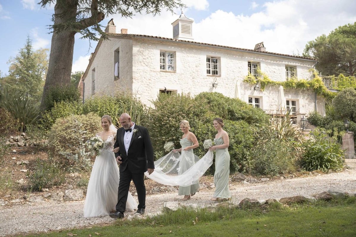 A bride is being escorted along a gravel path by a man in a black tuxedo with bow tie, likely her father, while two bridesmaids walk closely behind holding up the train of her cathedral-length lace-edged veil. The bride wears a white ballgown with a lace illusion bodice and carries a loose bouquet of white blooms and greenery; the two bridesmaids wear sage green satin slip dresses and one carries a small bunch of white gypsophila. The setting is outdoor, in front of a two-story whitewashed stone French country house or mas with terracotta roof tiles, climbing vines, and manicured garden borders. This is a wide shot captured during the processional walk to the ceremony, with a classic, understated styling palette of white, sage green, and black. Potential venue feature image.