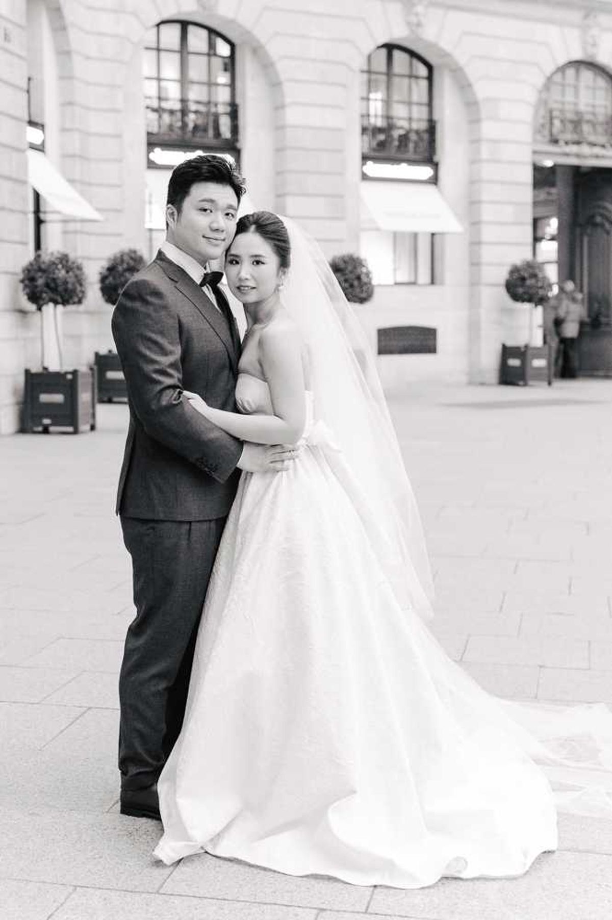 This is a black-and-white couple portrait taken outdoors in front of a grand Haussmann-style building with arched windows, wrought iron balconies, and topiary trees flanking the entrance, consistent with a Parisian plaza setting. The groom stands behind the bride with his arms wrapped around her waist; he wears a dark suit with a tie, while the bride wears a strapless ballgown with a full skirt, a long cathedral-length train, and a long veil worn down her back. Both face the camera with soft, composed expressions. The image has bright highlights and mid-range contrast with the bride's gown and veil rendered in bright whites against the light stone pavement. The composition is a full-length portrait shot with the architectural facade softly blurred in the background.