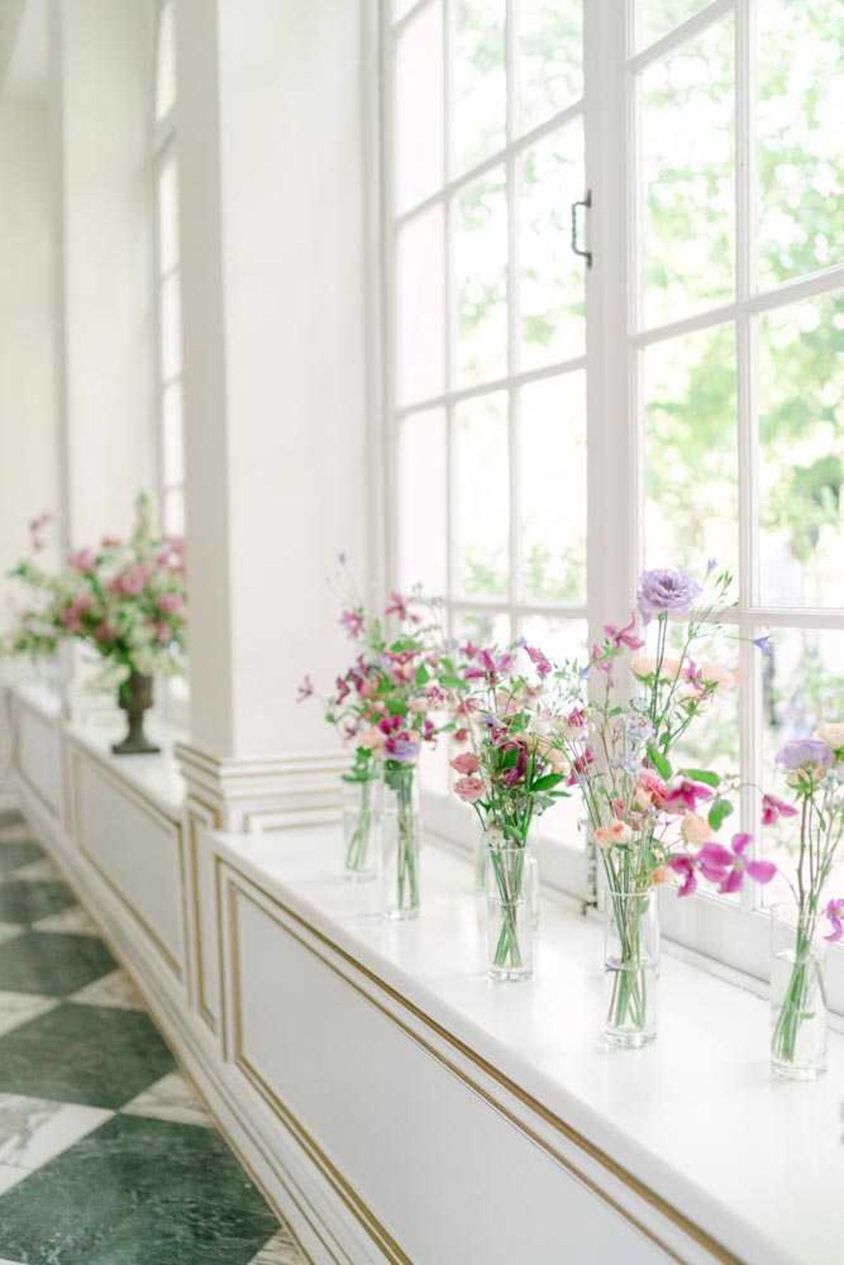 Detail shot of floral decor arranged along a window ledge inside a classic French venue, featuring multiple clear glass bud vases filled with loose, garden-style arrangements of pink sweet peas, blush ranunculus, lavender lisianthus, magenta garden roses, and trailing greenery. The vases are grouped in clusters of varying heights along the white window sill, with a larger urn-style arrangement visible further down the ledge. The interior space features white paneling with gold trim detailing and a green-and-white checkered marble floor, suggesting a chateau or orangerie setting. Natural light streams through tall white-framed French windows, creating a soft, bright atmosphere. Potential venue feature image.