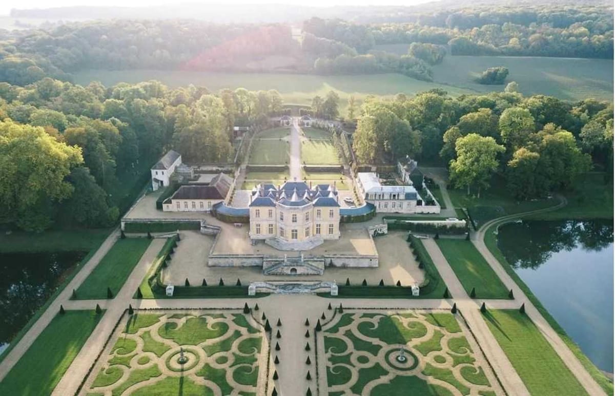 Aerial drone shot of a French classical chateau and its formal grounds, with no wedding activity or people visible. The chateau features pale stone facades, blue-grey slate mansard roofs with conical turrets, and symmetrical wings extending to either side. In the foreground, an ornate French parterre garden is laid out with scrollwork hedging patterns, topiary cones, and gravel pathways arranged in precise geometric designs around two small fountains. A central grand staircase leads from the parterre up to the chateau's entrance. Beyond the chateau, formal walled gardens with a central allée extend toward a wooded backdrop. A lake or ornamental pond is visible to the right of the property. The image is taken in soft early morning or late afternoon light with a slight warm lens flare. Potential venue feature image.