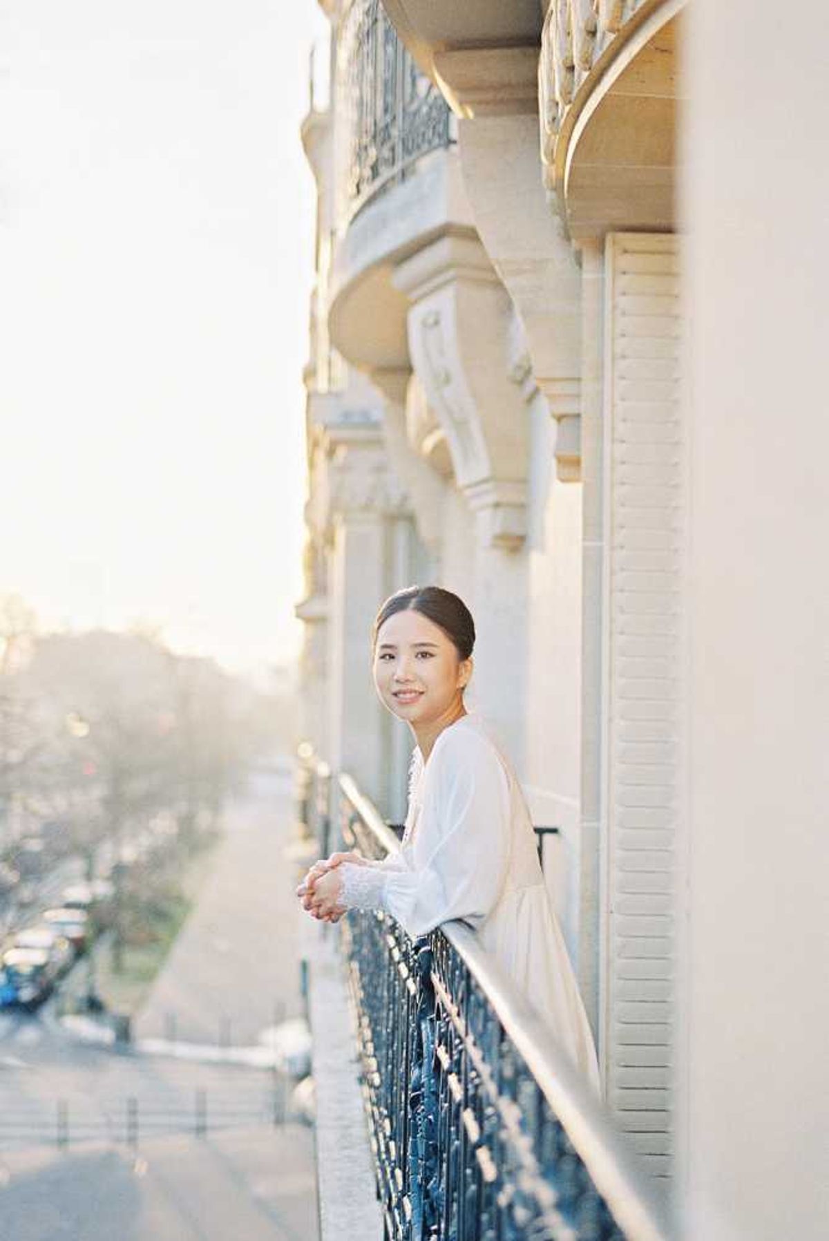A bride stands on an ornate wrought-iron balcony of a classic Haussmann-style building, leaning on the railing and looking back toward the camera with a relaxed smile. She wears a white long-sleeve bridal top or gown with delicate ruffle or lace cuff detailing, and her dark hair is pulled back in a low updo. The shot is taken during golden hour, bathing the cream stone facade and repeating arched balconies in warm, soft light. This is a portrait composition with a shallow depth of field, with a street and parked cars visible in the soft-focus background below.