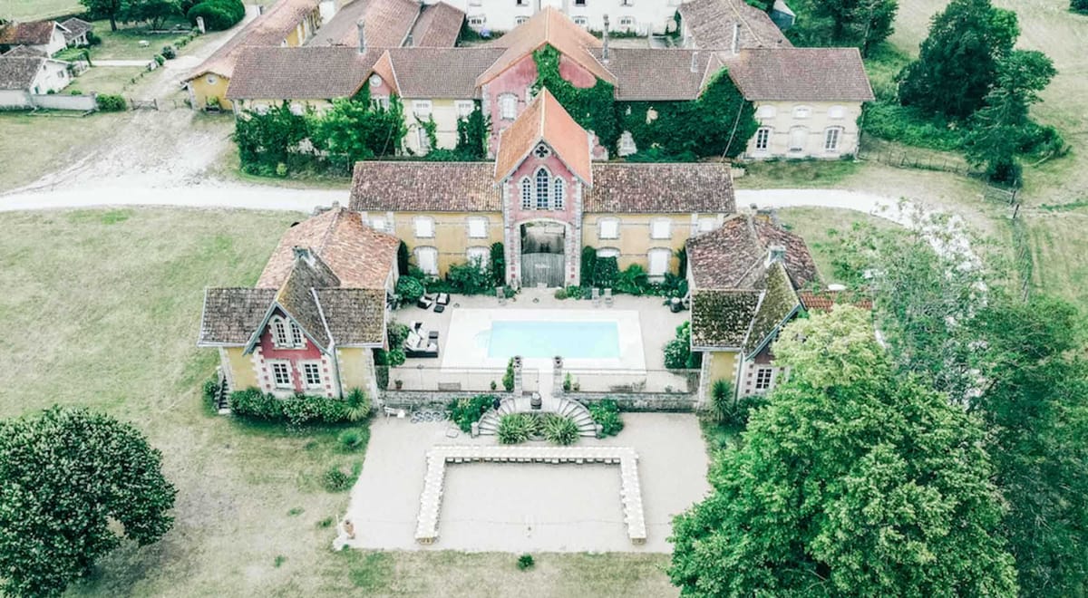 An aerial drone shot of a French country estate venue, showing the full property layout from above. The complex features a central building with a distinctive Gothic-arched gateway entrance in red brick, flanked by two symmetrical pavilions with terracotta-tiled roofs, and a rectangular swimming pool in the central courtyard. In the foreground, a ceremony space is visible with rows of white chairs arranged in a U-shape on a gravel terrace, connected to a formal garden area with a circular ornamental feature. The surrounding grounds include open fields, mature trees, and additional outbuildings with ochre-yellow rendered facades visible in the background. Potential venue feature image.