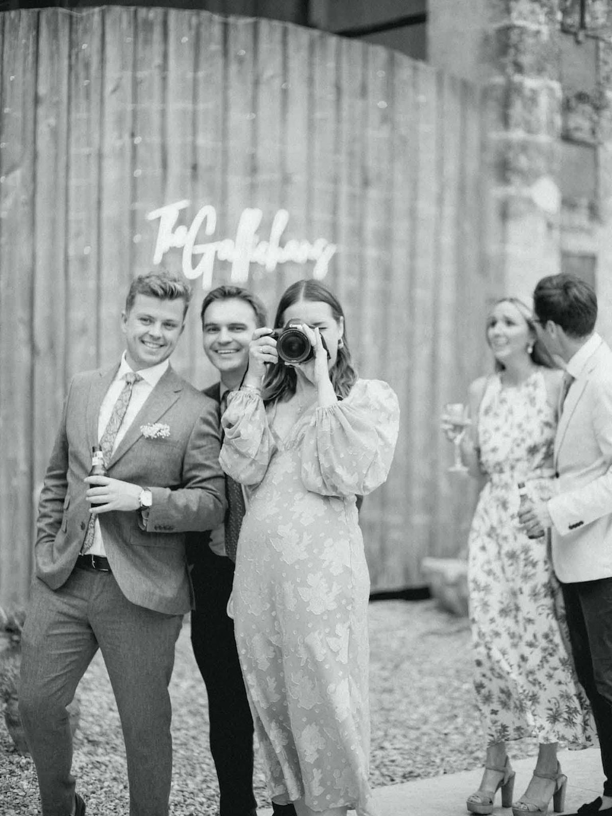 A black-and-white candid shot taken during what appears to be a cocktail hour or outdoor reception, with four guests gathered in front of a rustic wooden backdrop bearing a cursive script sign reading 'The Goldbergs' (or similar surname). In the foreground, a woman in a puff-sleeved midi dress with a textured floral pattern raises a DSLR camera toward the photographer, while two men in suits — one in a checked or herringbone suit with a floral tie and pocket square, holding a bottle — smile at the camera. To the right, a woman in a floral-print midi dress holds a wine glass and converses with a man in a light-colored blazer. The image has soft, slightly hazy tones consistent with film photography, with good contrast between the light clothing and the darker wooden backdrop behind them.