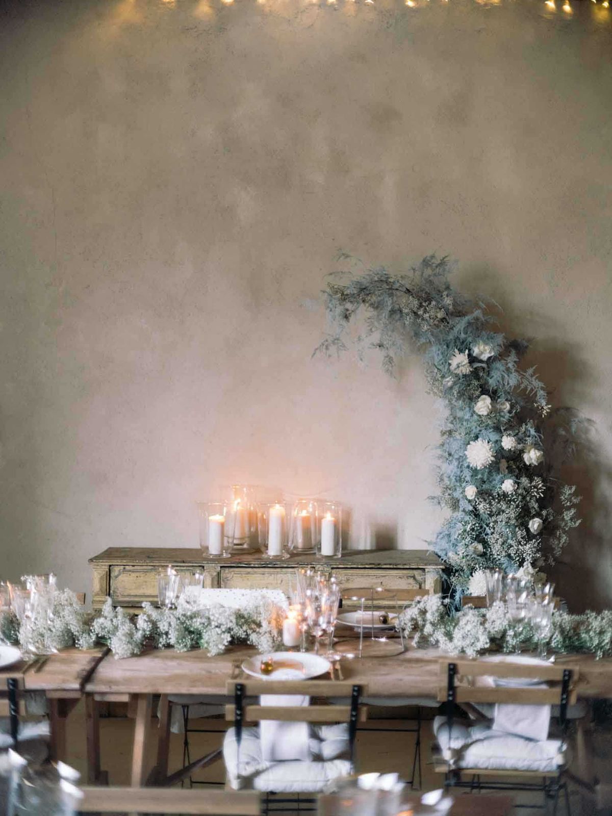 A detail shot of an indoor wedding reception table setup with no guests present. The long wooden farm table is dressed with a runner of white dried baby's breath interspersed with fairy lights, clear glassware, white plates, and white linen napkins on dark metal chairs. Behind the table sits a distressed antique sideboard topped with a cluster of white pillar candles in glass hurricane holders. To the right, a large crescent-shaped floral installation rises from floor level, composed of dusty blue-green foliage — including what appears to be eucalyptus and dried fern — accented with white garden roses and white pompom blooms. A strand of fairy lights runs along the top of the plastered wall, adding warm ambient light to the overall cool, muted color palette. The styling theme is romantic and organic with a cool blue-grey and white color scheme.