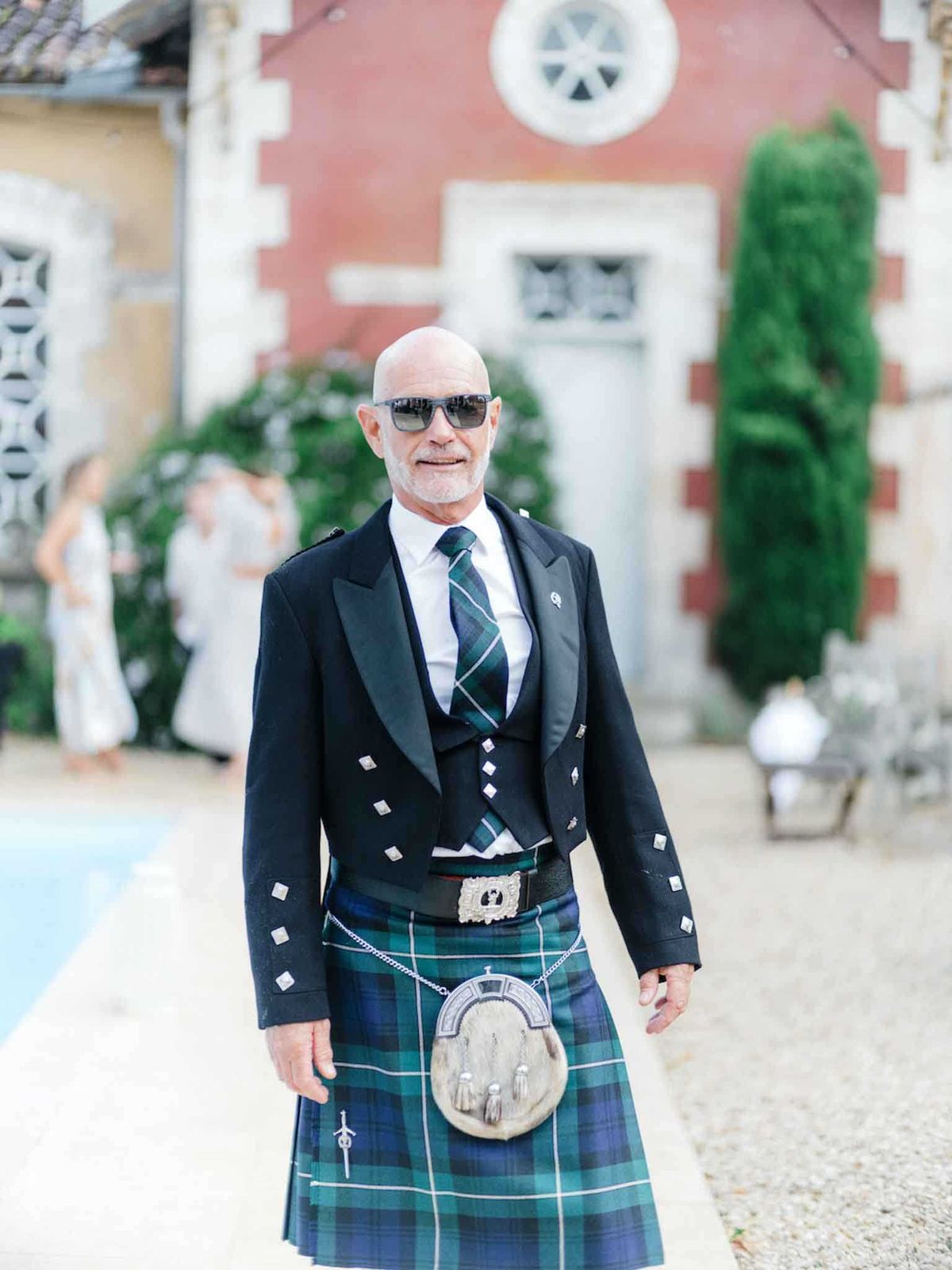 A male wedding guest poses for a portrait outdoors at what appears to be a French chateau property, wearing full traditional Scottish Highland dress including a green and navy blue tartan kilt, a black Prince Charlie jacket with silver buttons, a matching tartan tie, white dress shirt, leather belt with ornate silver buckle, and a fur sporran with silver hardware. He is bald, wearing dark sunglasses, and smiling at the camera. In the soft-focus background, several guests in light-colored attire are visible near what appears to be a pool, alongside a red-brick chateau building with stone quoins and a circular window detail. The image is a mid-length portrait shot with shallow depth of field.