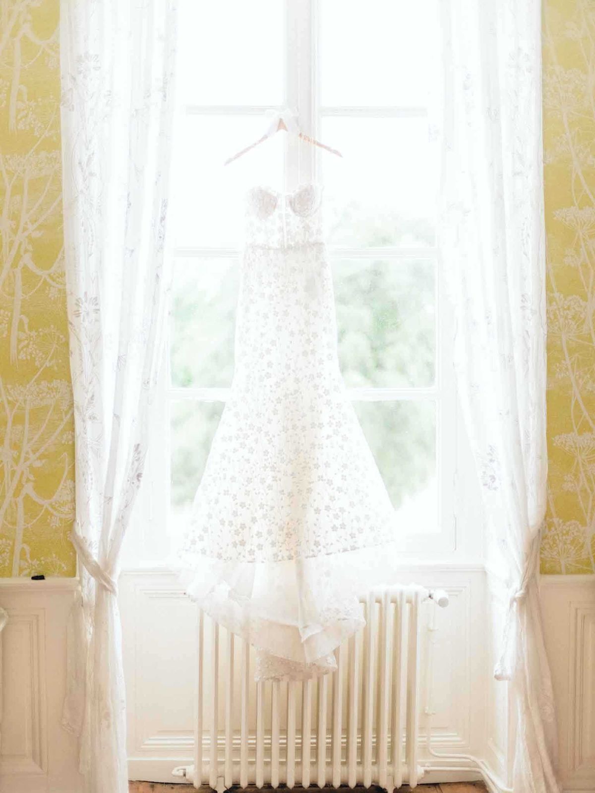 A getting-ready detail shot of a white strapless wedding dress hanging from a pale pink hanger in front of a tall French window, backlit by natural light. The dress features an all-over floral lace pattern across the bodice and skirt, with a fitted silhouette and what appears to be a tulle or lace hem at the bottom. The room walls are covered in yellow-gold botanical-patterned wallpaper with white floral motifs, and sheer white lace curtains frame the window on either side. A white cast-iron radiator is visible beneath the window, adding to the classic French interior feel of the bridal preparation space.