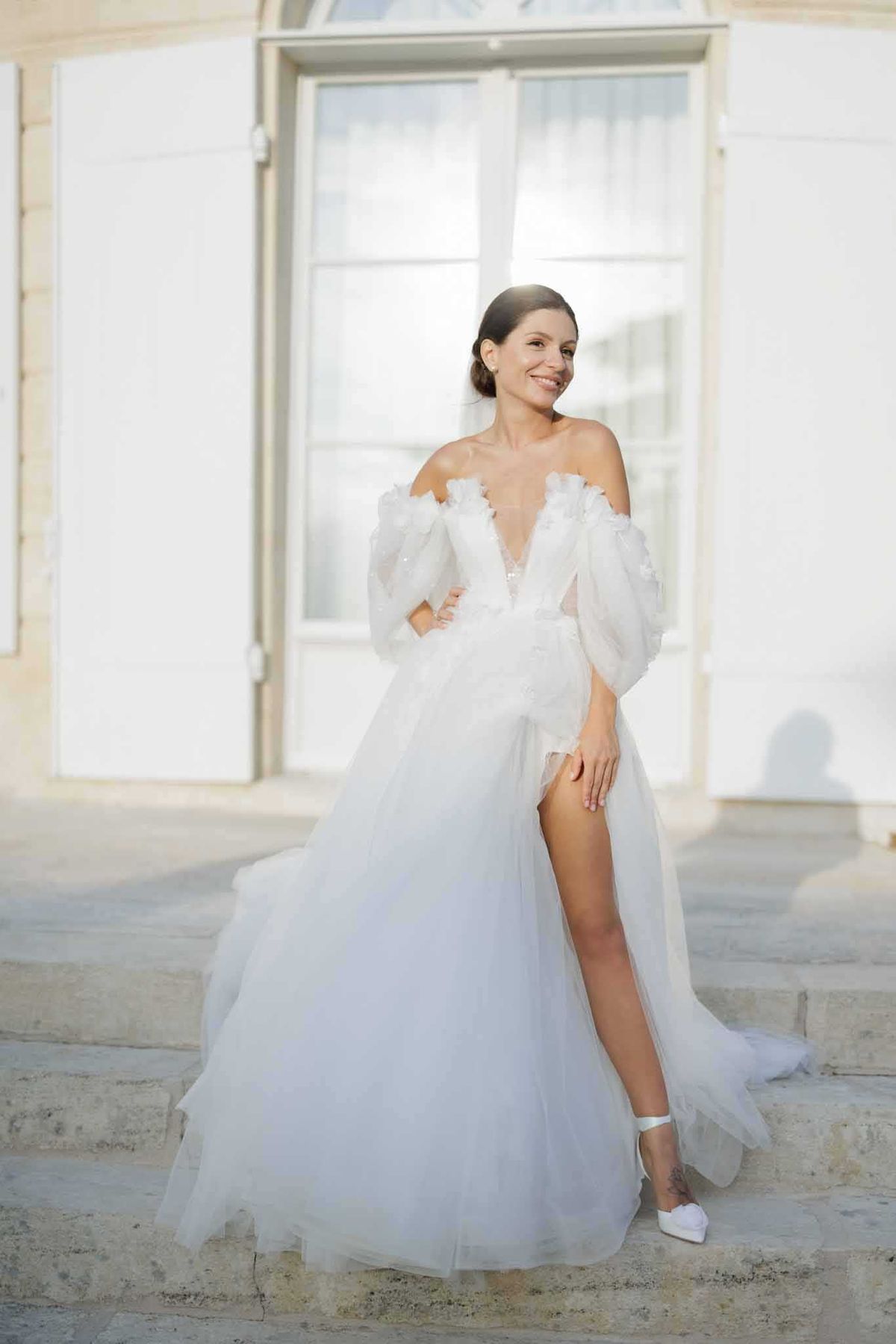 A bridal portrait taken outdoors on the stone steps of what appears to be a French chateau or manor house, with white shuttered arched windows visible in the background. The bride stands alone, smiling off-camera, wearing an off-the-shoulder white tulle A-line gown with a deep V-neckline, 3D floral appliqué details along the bodice and sleeves, billowing detached long sleeves, and a high front slit revealing white ankle-strap heeled sandals. Her dark hair is styled in a low updo, and the overall aesthetic is modern romantic with a fashion-forward edge. The shot is a full-length portrait with soft, bright natural light creating a clean, airy feel.
