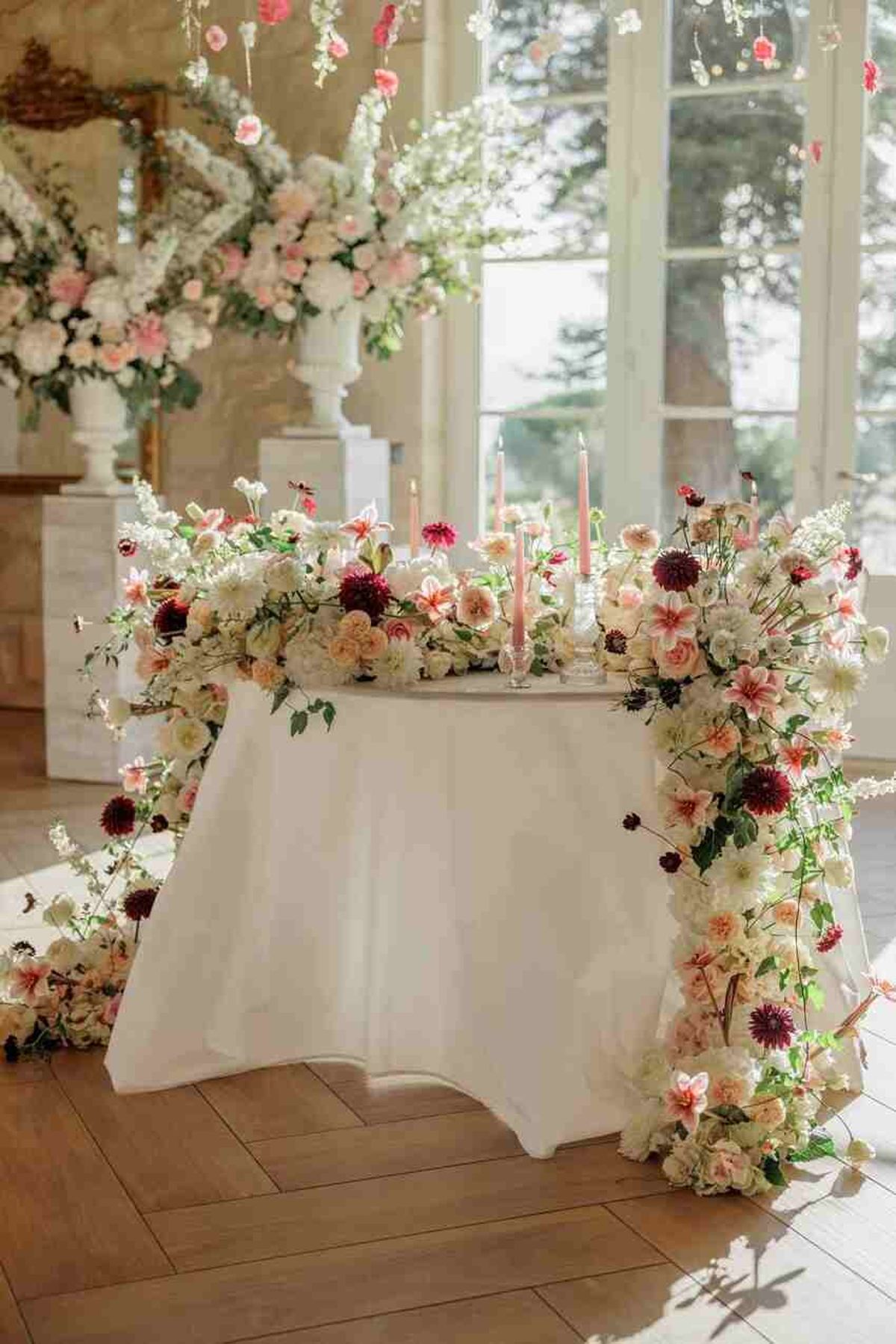 A detailed reception decor shot of a sweetheart table covered in a white floor-length linen, set inside a bright classical interior with tall French windows, herringbone wood flooring, and ornate architectural columns. The table is heavily decorated with a cascading floral installation featuring cream and ivory roses, blush and peach dahlias, burgundy dahlias, coral garden roses, white stock flowers, and trailing greenery that spills down both sides of the table and pools onto the floor. Three blush pink taper candles in glass candlestick holders are placed along the table surface. In the background, additional large white urn arrangements and overhead hanging floral installations continue the same color palette of white, blush, pink, and deep burgundy. The overall styling theme is romantic and garden-inspired with a rich, full floral aesthetic. Wide shot with no people present.