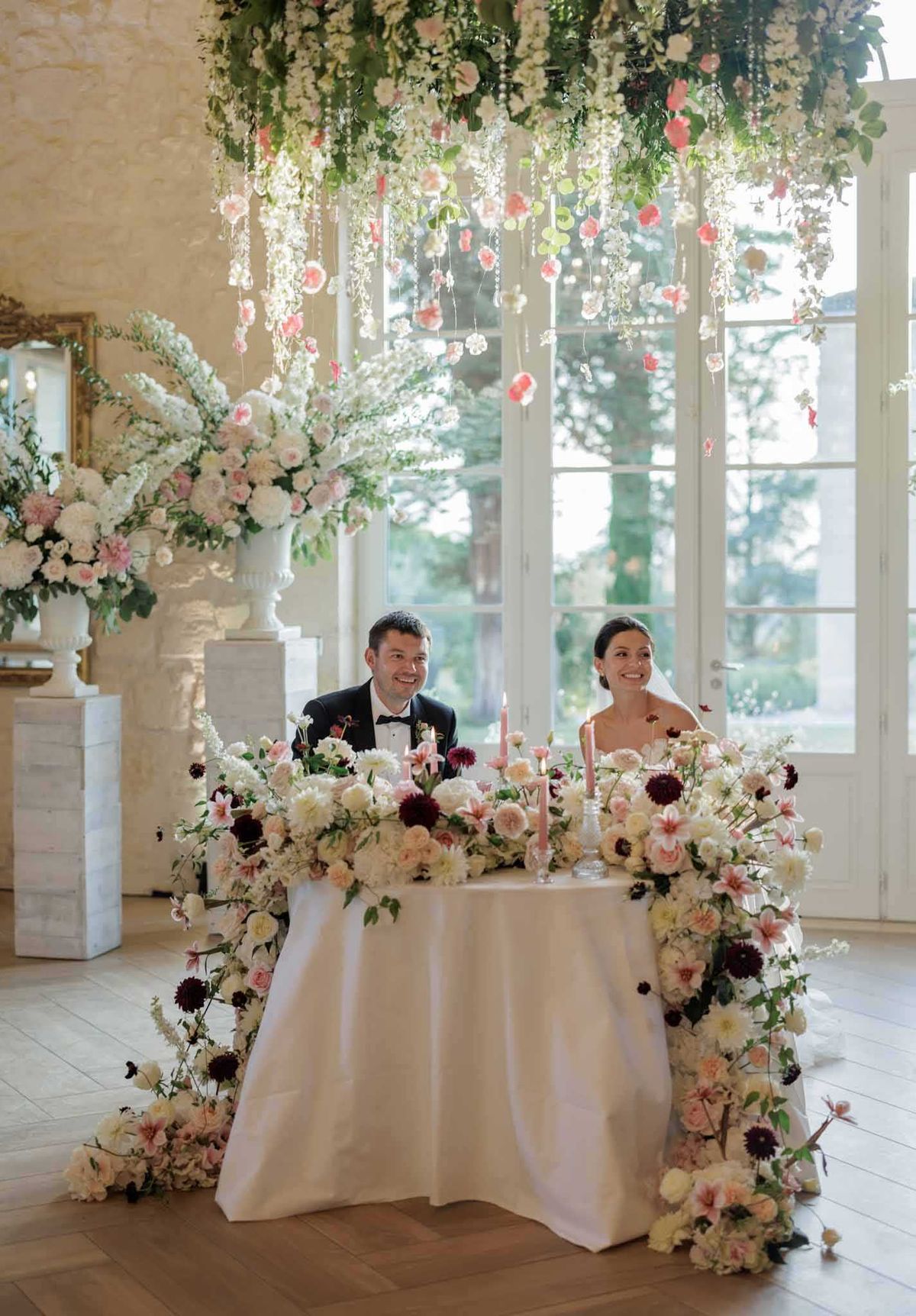 The bride and groom are seated at a sweetheart table during an indoor reception in what appears to be a French chateau ballroom with warm stone walls, tall French doors, and parquet flooring. The table is draped in a white linen and completely surrounded by an abundant floral installation that cascades from the tabletop down to the floor, featuring ivory garden roses, blush and coral dahlias, burgundy dahlias, white delphinium, and trailing greenery. Pink taper candles are placed among the florals on the table. Above the couple, a large hanging floral chandelier displays white and coral-pink blooms, crystal or pearl strands, and trailing greenery. A large white urn arrangement with a similar blush and white palette sits on a pedestal to the left. The groom wears a black tuxedo with a bow tie, and the bride wears a strapless white gown with a veil, her dark hair styled up. A gilded mirror is partially visible on the left wall. The shot is a wide portrait framing both the couple and the full floral installation.
