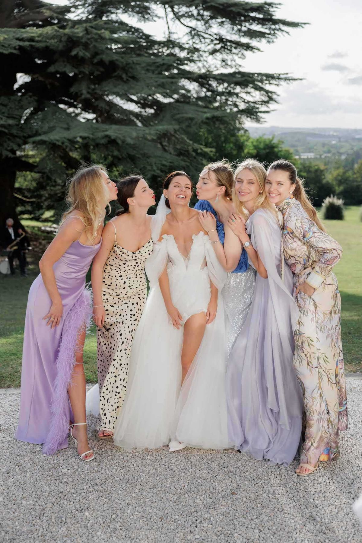 A candid group portrait of a bride and five bridesmaids/female guests posing outdoors on a gravel path, set against manicured grounds with a large cedar tree and rolling landscape in the background. The bride wears an off-the-shoulder white ballgown with a tulle skirt and veil, laughing as the women around her lean in to kiss her cheeks. The five guests wear a mix of mismatched dress styles — a lilac satin gown with feather trim and a thigh slit, a black-and-white dalmatian-print slip dress, a cobalt blue long-sleeved sequined dress, a lavender flowing chiffon gown, and a floral-printed long-sleeve maxi dress — suggesting a non-uniform bridesmaids look with a soft purple and pastel color theme. The shot is a full-length portrait with a slightly shallow depth of field, taken in natural daylight.