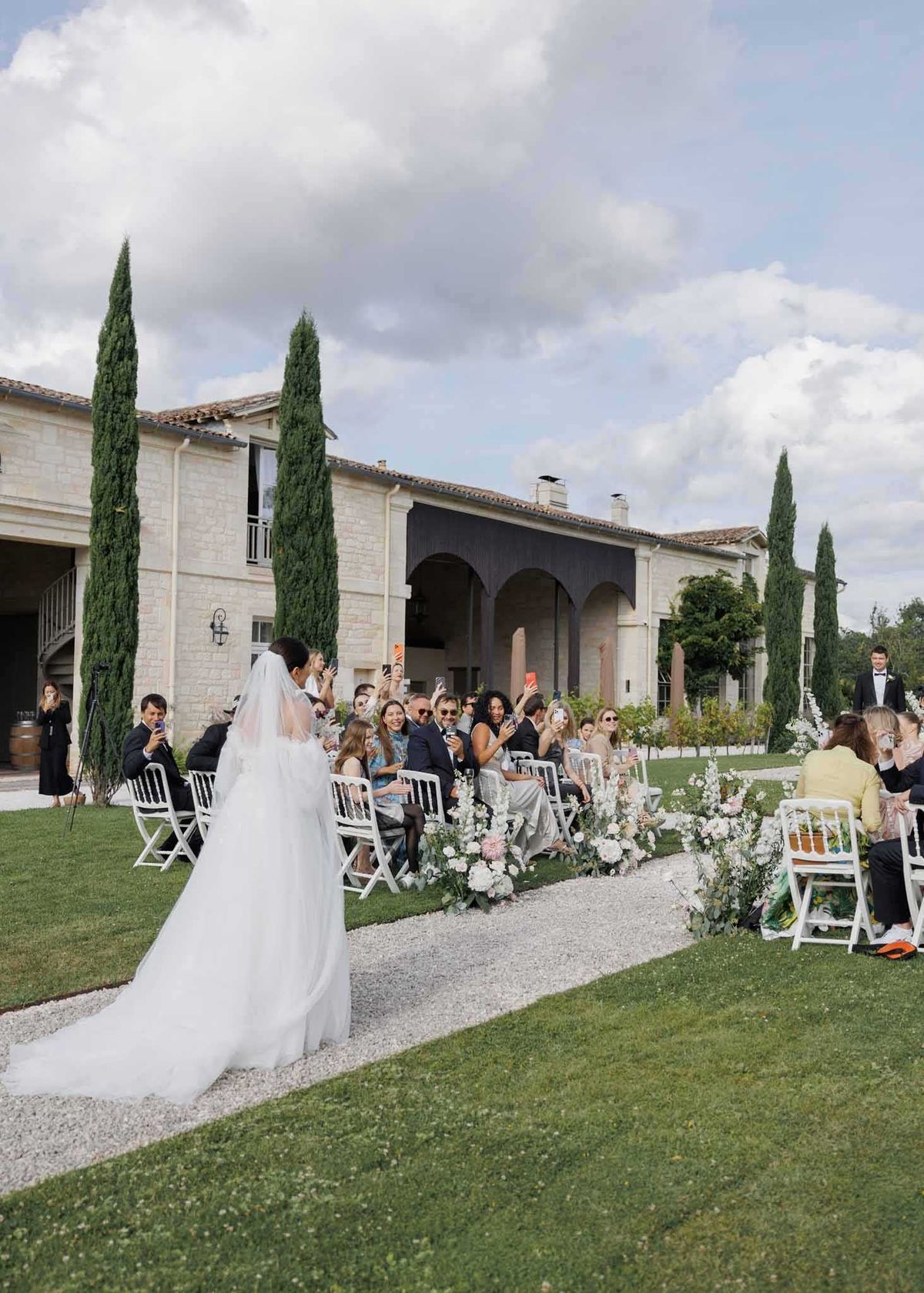 An outdoor wedding ceremony is underway at a French estate, with the bride walking down a gravel aisle toward the altar. The bride wears a white ball gown with a long cathedral-length veil and a trailing skirt. Approximately 30 guests are seated on white folding chairs arranged in rows on either side of the aisle, many holding up phones to photograph the procession. The aisle is lined with low floral arrangements featuring blush pink dahlias, white blooms, and trailing greenery. The ceremony takes place on a manicured lawn in front of a large limestone chateau with arched colonnades, with tall Italian cypress trees framing the building. The groom is visible standing at the far end of the aisle in a black tuxedo. This is a wide shot taken from behind and to the side of the bride. Potential venue feature image.