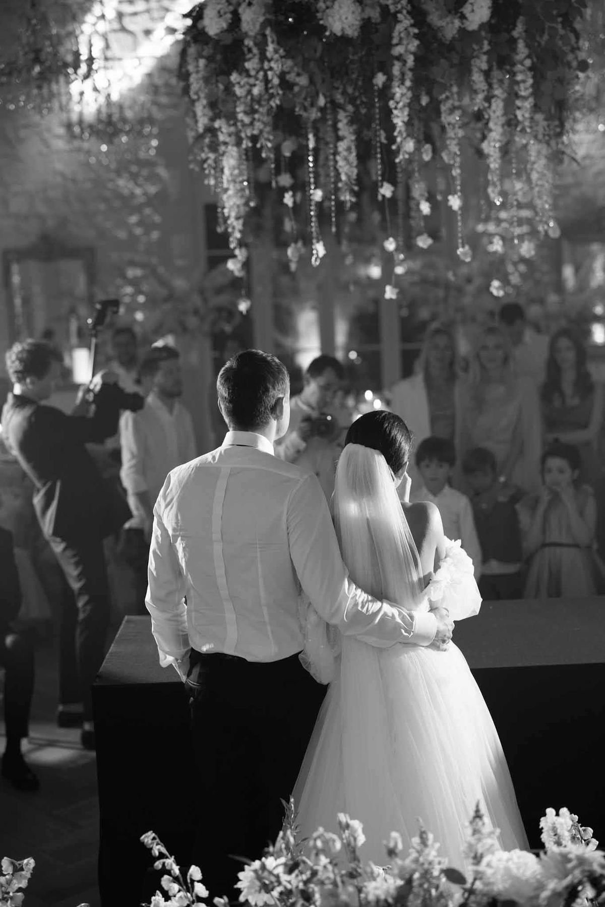 Black-and-white portrait-style shot of a bride and groom standing at the altar during an indoor wedding ceremony, photographed from behind. The groom wears a white dress shirt and dark trousers, while the bride wears a full tulle gown with a long veil and holds a bouquet. The ceremony space features an elaborate overhead installation of cascading florals and hanging crystal or bead strands, creating a dense canopy above the couple. Floral arrangements are also visible in the foreground at the base of the altar. An officiant stands facing the couple in the middle ground, with approximately 15–20 seated and standing guests visible on both sides in the background. The interior space appears to have stone walls with warm uplighting visible in the mid-tones, suggesting a barn or chapel venue. The image has soft, warm contrast with bright highlights on the bride's veil and gown against the darker background.