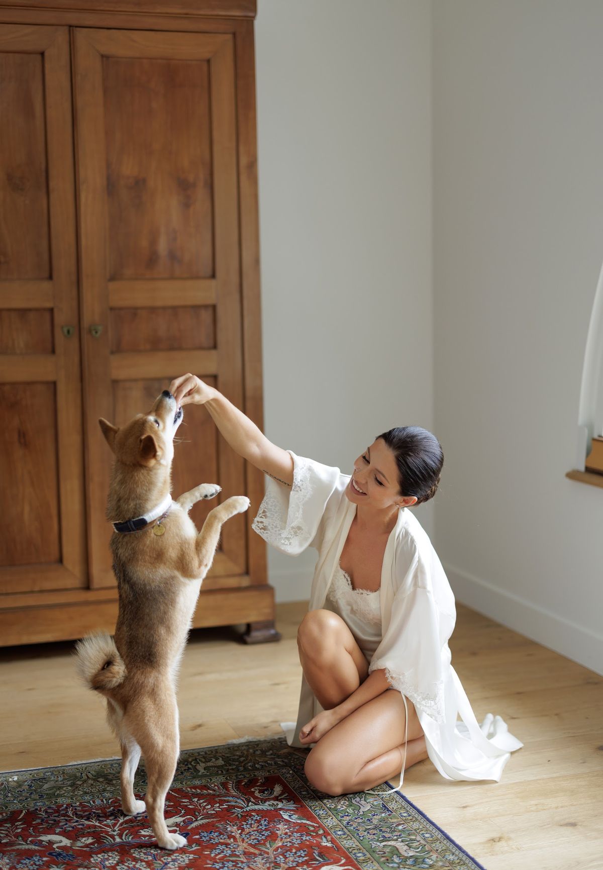 A getting-ready moment captured indoors, showing a bride kneeling on a red patterned area rug on a light wood floor, playing with a Shiba Inu dog that is standing on its hind legs reaching for a treat. The bride is wearing a white lace-trimmed bridal robe over white lingerie, with her dark hair styled up, and is smiling down at the dog. The room features a large antique dark wood armoire and neutral white walls, suggesting a classic French interior. The shot is a full-length portrait with a candid, relaxed feel.