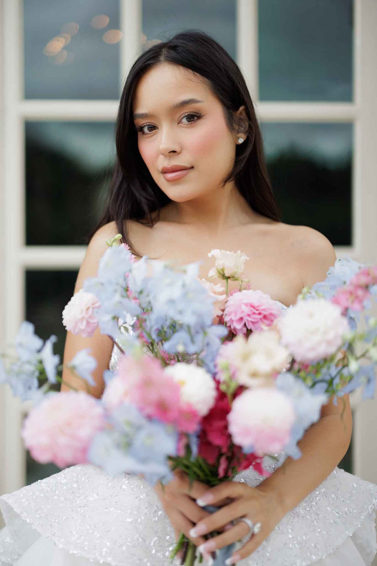 A close-up bridal portrait of a bride with long dark hair worn loose, photographed outdoors in front of large white-framed windows. She wears a strapless or off-shoulder white gown with allover sequin or beaded embellishment, and small diamond stud earrings. She holds a voluminous bouquet composed of powder blue delphiniums, pink and hot pink carnations or peonies, blush lisianthus, and white blooms, creating a soft pastel palette. The composition is a medium portrait shot with shallow depth of field, keeping the bride sharp against a softly blurred architectural background.