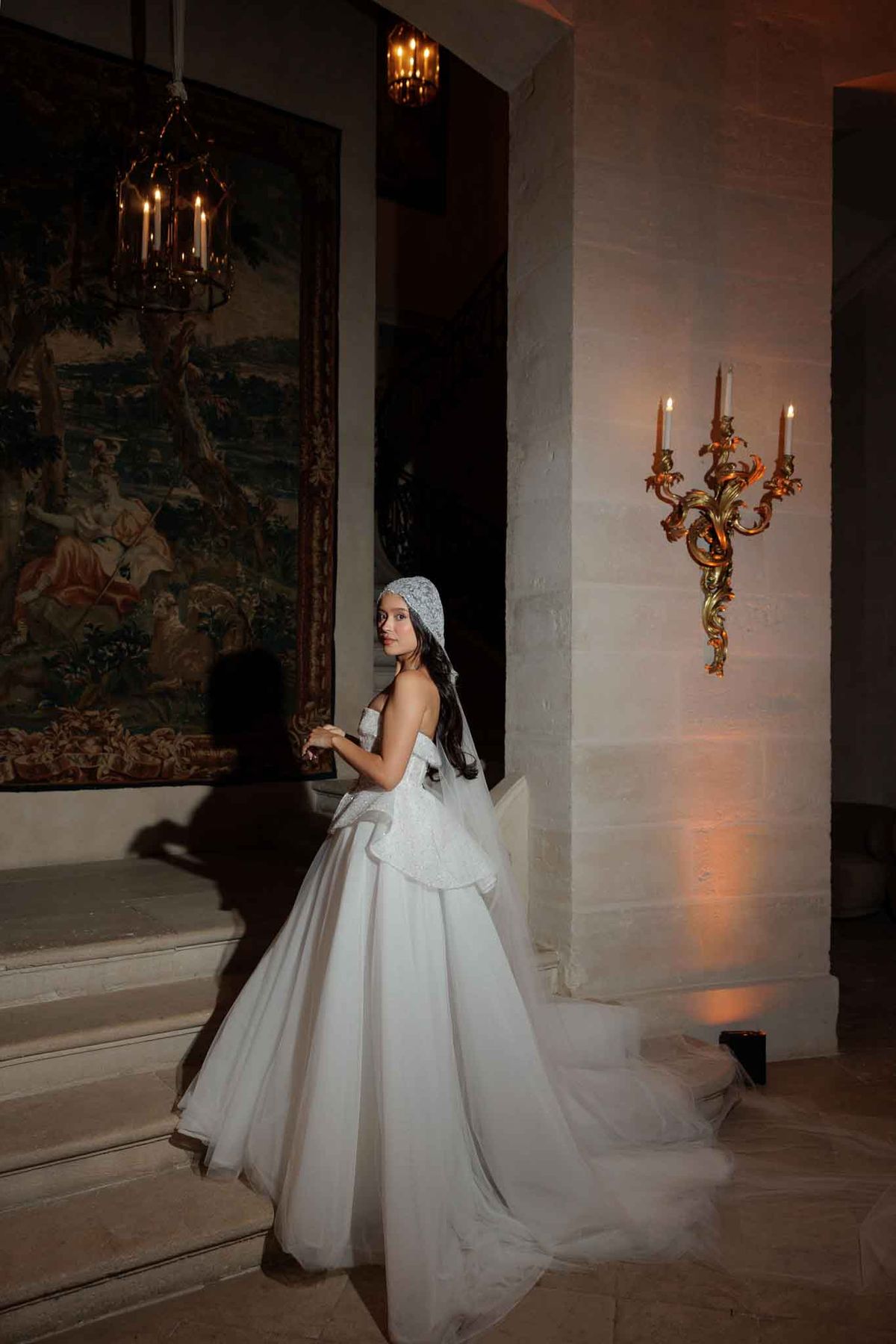 A solo bridal portrait taken indoors in what appears to be a French chateau, with the bride standing on stone steps and glancing back over her shoulder toward the camera. She wears a white strapless ball gown with a structured peplum bodice featuring textured or embroidered detailing, a full tulle skirt with a long train, and a lace or beaded juliet cap with a long veil trailing down her back. The setting is lit with warm amber uplighting, a gold ornate candelabra wall sconce with lit candles, and a hanging brass lantern with candle-style bulbs. A large antique tapestry with figurative scenes in earthy tones hangs on the wall to the left. The overall styling aesthetic is classic and formal with baroque-inspired decor. Full-length portrait shot with strong directional lighting creating dramatic shadows.