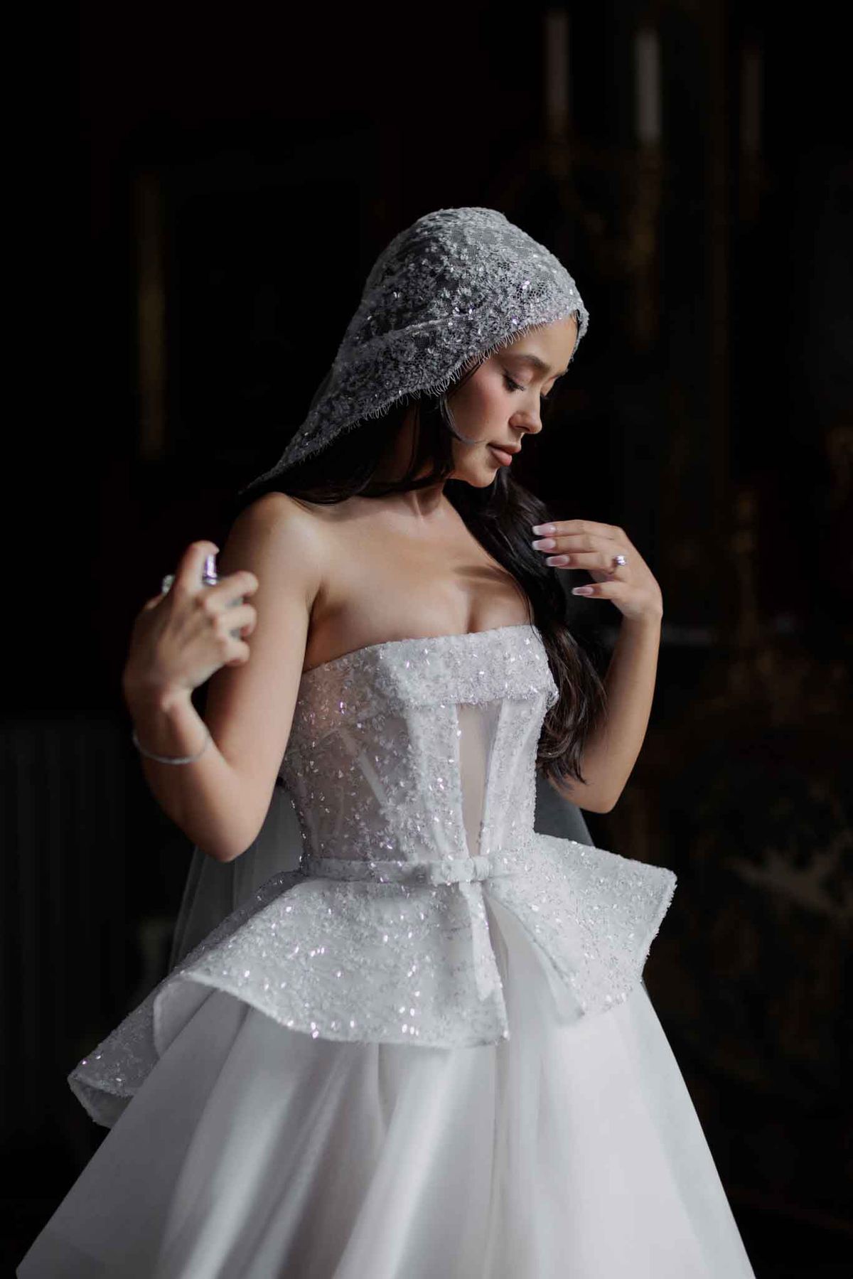 A bridal portrait of a single bride photographed against a very dark, almost black background, creating strong chiaroscuro lighting that highlights her face and gown. She wears a strapless white ball gown with a heavily embellished sequined and beaded corset bodice featuring a structured peplum with a large bow detail at the waist, over a full tulle skirt. Her head is covered with a heavily beaded and sequined juliet-style cap veil that drapes down over her dark hair. She looks downward with her hands raised toward her shoulders, one hand appearing to hold a small perfume bottle. Her nails are painted in a soft lavender shade and she wears a delicate bracelet. The overall styling is modern and highly embellished with a structured, architectural aesthetic. Close-up portrait shot from approximately the waist up.