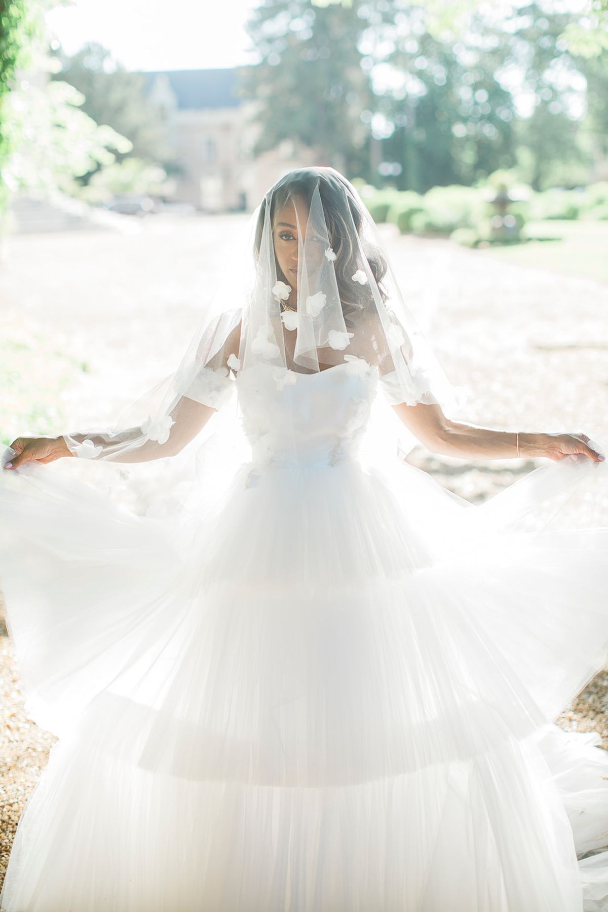 A bridal portrait taken outdoors on a gravel path, with a chateau building and manicured garden visible in the softly blurred background. The bride is facing the camera while spreading her voluminous white tulle ball gown skirt wide with both arms outstretched, and a two-tier veil adorned with scattered white 3D floral appliqués drapes over her face and shoulders. She wears a structured sweetheart-neckline bodice and gold statement earrings, with loose wavy hair beneath the veil. The image is shot in bright, high-key natural light with a slightly overexposed quality, giving the white tulle layers a luminous, airy appearance, captured in a medium full-length portrait composition.
