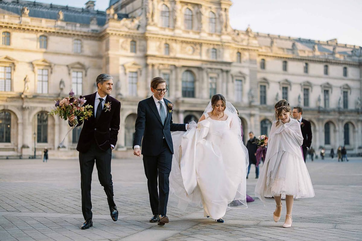 A wedding portrait session taking place in the courtyard of the Louvre museum in Paris, with the classical stone facade visible in the background. The bride wears a white off-the-shoulder ball gown with a long veil and holds the groom's hand as they walk across the cobblestone plaza; the groom is dressed in a navy suit with a grey tie and a floral boutonniere. To the left, a male guest in a burgundy velvet blazer and dark trousers carries a loose, garden-style bouquet of mauve, burgundy, and dusty pink blooms with dark foliage; to the right, a young flower girl in a white tea-length dress and floral crown wears a white wrap shawl. Additional members of the wedding party are visible walking behind the group in the mid-ground. Wide candid-style shot taken at golden hour with warm directional light.