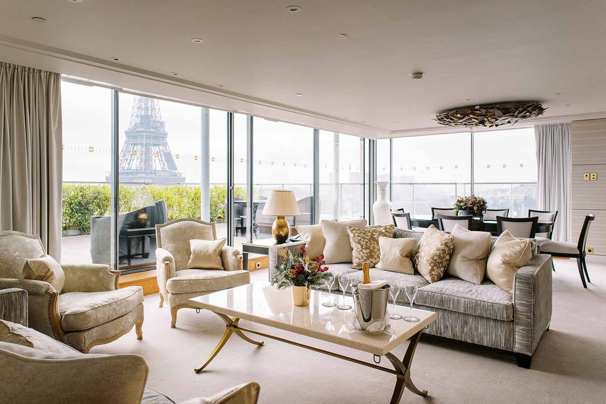 Interior wide shot of a high-end Paris hotel suite or penthouse living room with direct views of the Eiffel Tower through floor-to-ceiling glass windows. The space features a neutral palette of champagne, ivory, and soft grey upholstered furniture including a large sofa, armchairs with carved gilt legs, and a marble-topped coffee table with gold X-frame legs. A champagne bottle in an ice bucket and several champagne flutes are arranged on the coffee table alongside a small holiday floral arrangement with red berries and greenery. A dining area with dark wood chairs and a floral centerpiece is visible in the background. The decor style is classic Parisian with gold accents, a sculptural ceiling fixture, and ivory drape curtains. No people are present. Potential venue feature image.