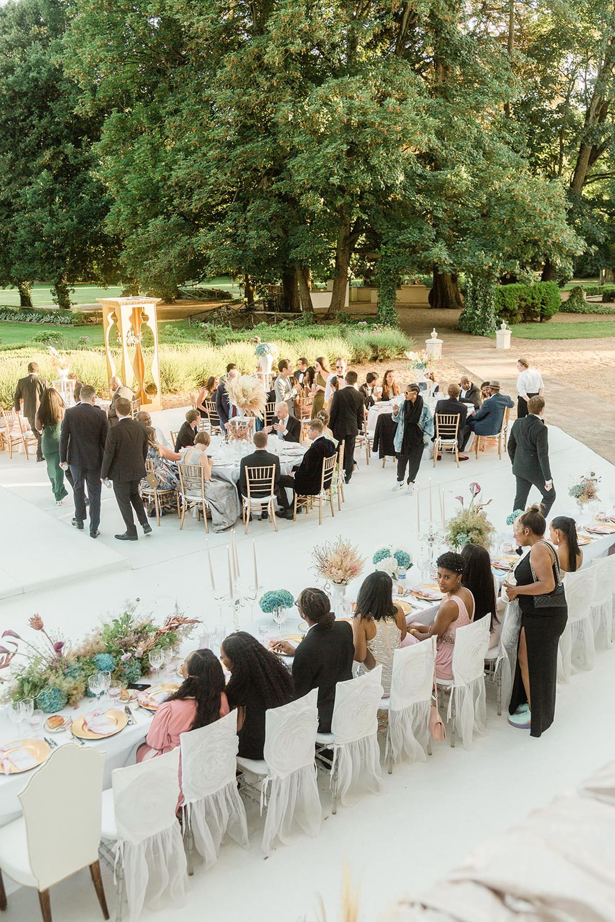 An outdoor wedding reception is underway on a large white-painted terrace surrounded by mature trees and manicured garden grounds. Approximately 50 or more guests are seated and mingling across a mix of long rectangular tables and round tables; the rectangular tables in the foreground are dressed with white ruffled chair covers and chiavari chairs, white linen runners, gold charger plates, tall white taper candles in silver candelabras, and centerpieces featuring teal-blue hydrangeas, dried pampas grass, burgundy proteas, and mixed dried botanicals in an earthy, bohemian palette. Round tables in the background use gold chiavari chairs without covers and similarly styled floral arrangements. A gold decorative structure, possibly a chuppah or decorative arch, is visible at the upper left. Guests are dressed in formal and semi-formal attire including black suits, gowns in blush pink, black, and silver tones. The shot is taken from an elevated angle, providing a wide overhead view of the full reception layout.