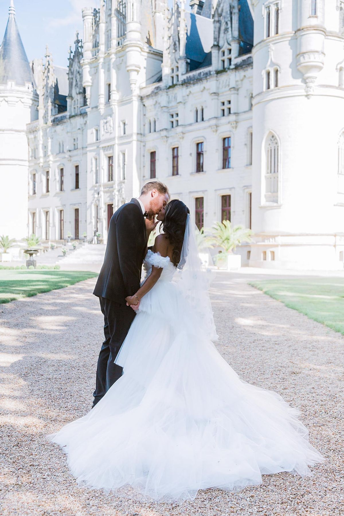 A couple portrait taken outdoors on a gravel path in front of a large French Gothic-style chateau with white stone facades, pointed turrets, and slate-blue rooftops. The bride and groom are leaning in to kiss, holding hands, centered in the frame with the chateau filling the background. The bride wears a strapless white ballgown with layered ruffled tulle skirt, an extended cathedral-length train spread across the gravel, and a mid-length veil; the groom wears a dark charcoal suit with a red tie. The image is shot as a full-length portrait with soft, bright natural light and a slightly overexposed background that draws focus to the couple. Potential venue feature image.