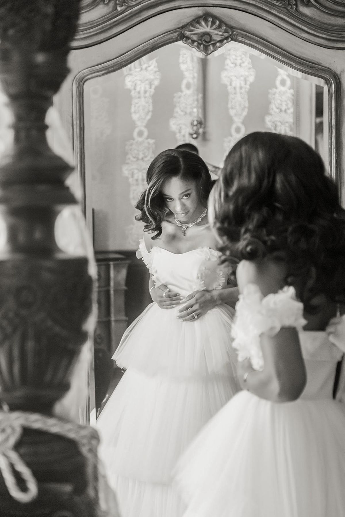 This black-and-white image captures a bride during the getting-ready phase, viewed from behind as she looks at her reflection in a large ornate mirror with carved gilded framing. The reflection shows her wearing a strapless ballgown with a tiered tulle skirt and floral appliqué detail along the bodice, paired with a statement necklace and her engagement ring visible on her hand. Her dark hair is styled in soft curls pinned up, and she gazes toward the camera with a subtle smile. The indoor setting features decorative wall paneling with raised plasterwork visible in the mirror's reflection, suggesting a classic chateau or grand period interior, and the composition uses a portrait angle with soft, bright highlights and deep shadows giving the image strong tonal contrast.