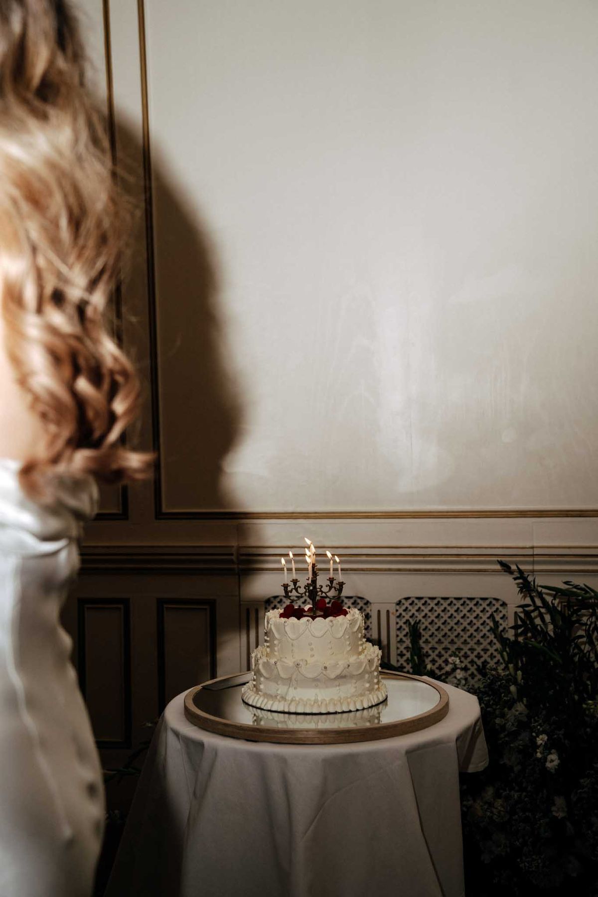 A cake cutting or cake reveal moment captured indoors, with the bride — visible only from behind and slightly blurred in the foreground — facing a single-tier white wedding cake displayed on a round table with a white linen and a wooden tray base. The cake features piped white frosting with scalloped detailing, deep red floral accents at the top, and a small ornate candelabra cake topper with five lit candles. The setting appears to be a classic interior room with cream paneled walls trimmed in gold molding, and dark green foliage greenery visible to the right. The bride has curly, honey-blonde hair and wears a white satin gown with a large back bow. The image is a medium-wide portrait shot with a moody, dimly lit atmosphere.