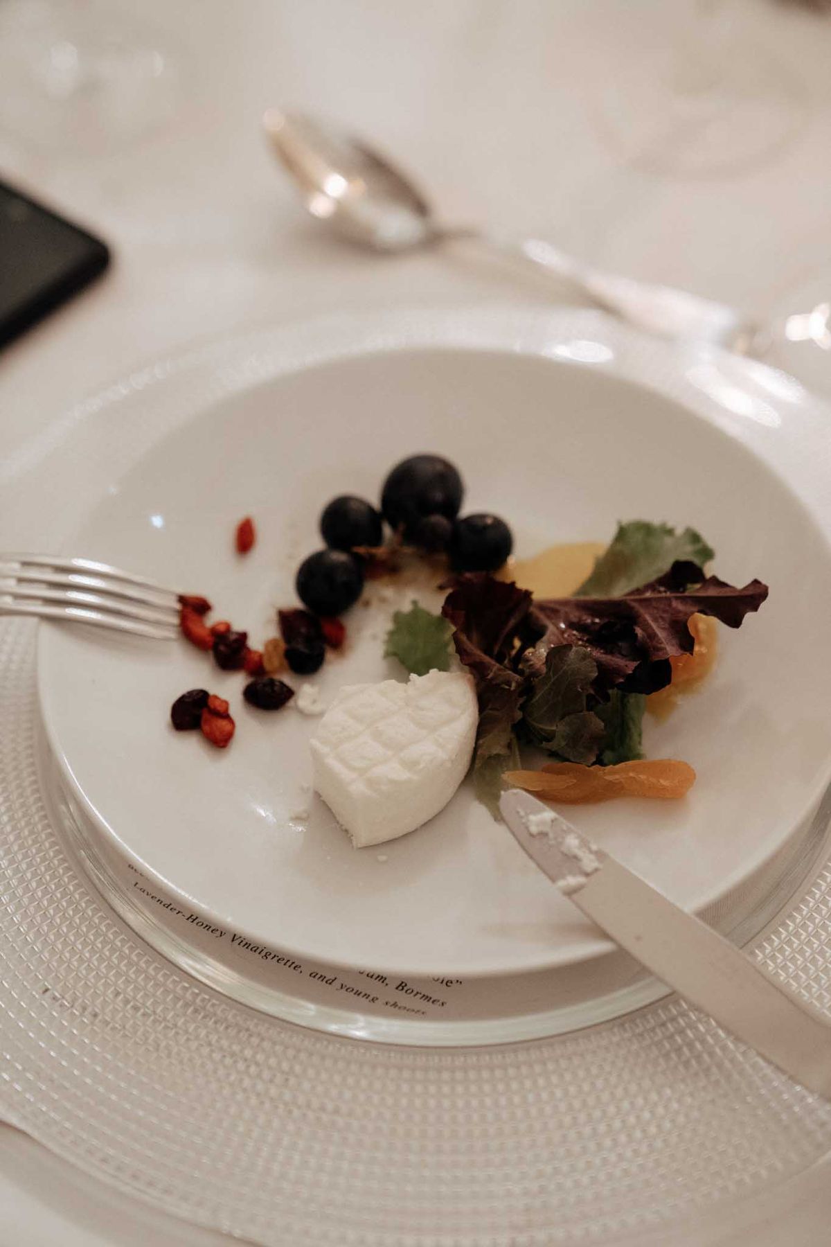 Close-up detail shot of a wedding reception dinner plate, partially eaten, resting on a white textured placemat. The white ceramic plate features printed text around its rim referencing 'Lavender-Honey Vinaigrette' and 'Bormes,' indicating a custom-printed menu plate used as a place setting. The dish includes a round portion of soft white cheese, fresh blueberries, mixed salad leaves in green and burgundy, dried apricot pieces, and small dried red berries or cranberries. Silver cutlery — a knife and fork — rest on the plate, and a spoon and wine glass are visible in the soft-focus background on a white linen tablecloth.