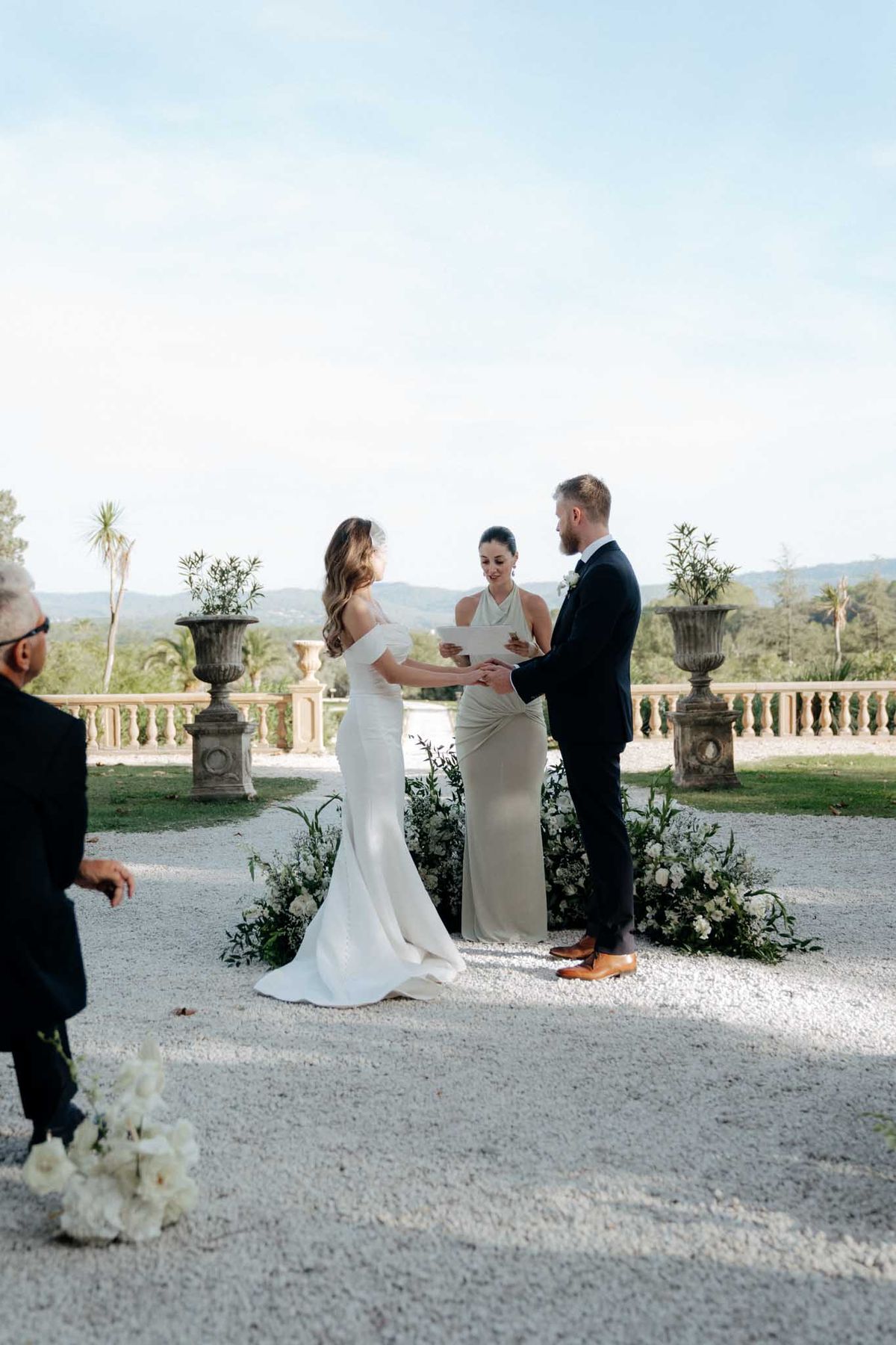 An outdoor wedding ceremony taking place on a gravel terrace, with a couple exchanging vows while holding hands facing each other. The bride wears an off-the-shoulder fitted ivory gown with a short veil, and the groom is dressed in a navy suit with a white boutonniere and tan Oxford shoes. A female officiant in a sage halter-neck column dress holds a paper and reads aloud between them. The ceremony backdrop features a stone balustrade terrace with large classical stone urns, and ground-level floral arrangements of white blooms and dark green foliage arc around the couple's feet. A guest in a dark suit is partially visible in the foreground left, alongside a cluster of white flowers. The overall styling is modern and minimal with a restrained white and green floral palette. Wide shot taken from a slight distance, capturing the full scene.