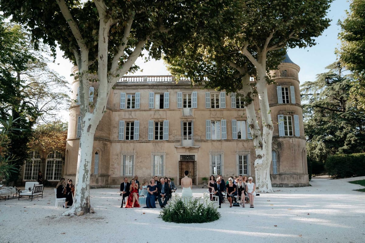 An outdoor wedding ceremony taking place on a gravel forecourt directly in front of a large French chateau featuring pale stone facades, pale blue shutters, rounded corner towers, and a balustraded roofline. Approximately 30–40 guests are seated in white chairs arranged in rows facing the chateau's entrance, where a bride in a backless white fitted gown stands at the altar area, which is decorated with a low ground-level arrangement of white florals and greenery. Guests are dressed in formal attire including dark suits, a red gown, and navy dresses; a musician playing violin is visible to the left near lounge seating. The composition is a wide shot taken from behind the seated guests, framing the chateau as the backdrop between two large plane trees. Potential venue feature image.