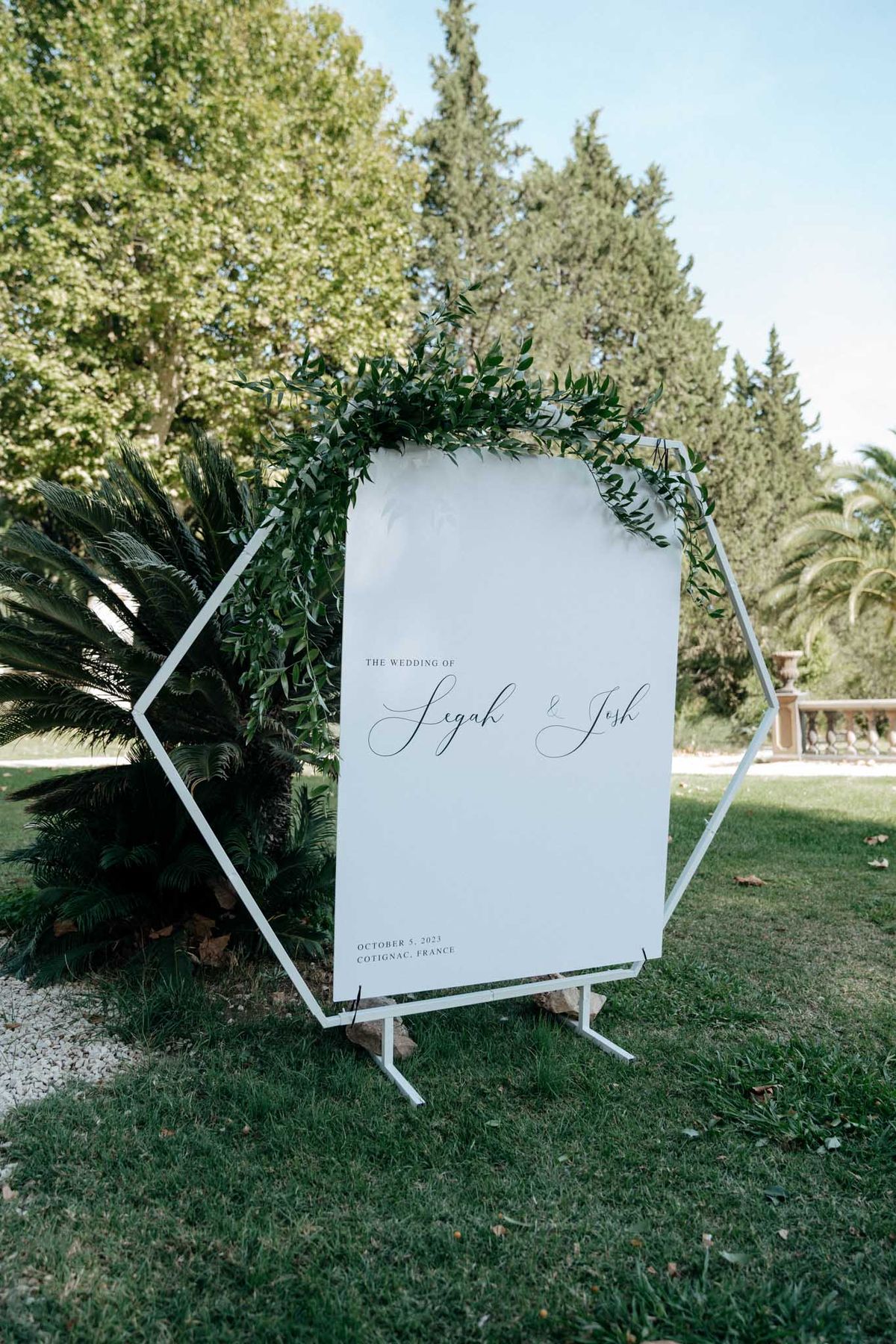 A wedding welcome sign displayed outdoors on a lawn in Cotignac, France, dated October 5, 2023. The sign reads 'The Wedding of Legah & Josh' in calligraphy script on a white board, mounted within a white geometric hexagonal metal frame stand adorned with a garland of dark green foliage along the top edge. The styling is modern and minimal, using a white-and-green color palette with clean lines. The shot is a medium-distance detail image centered on the signage.