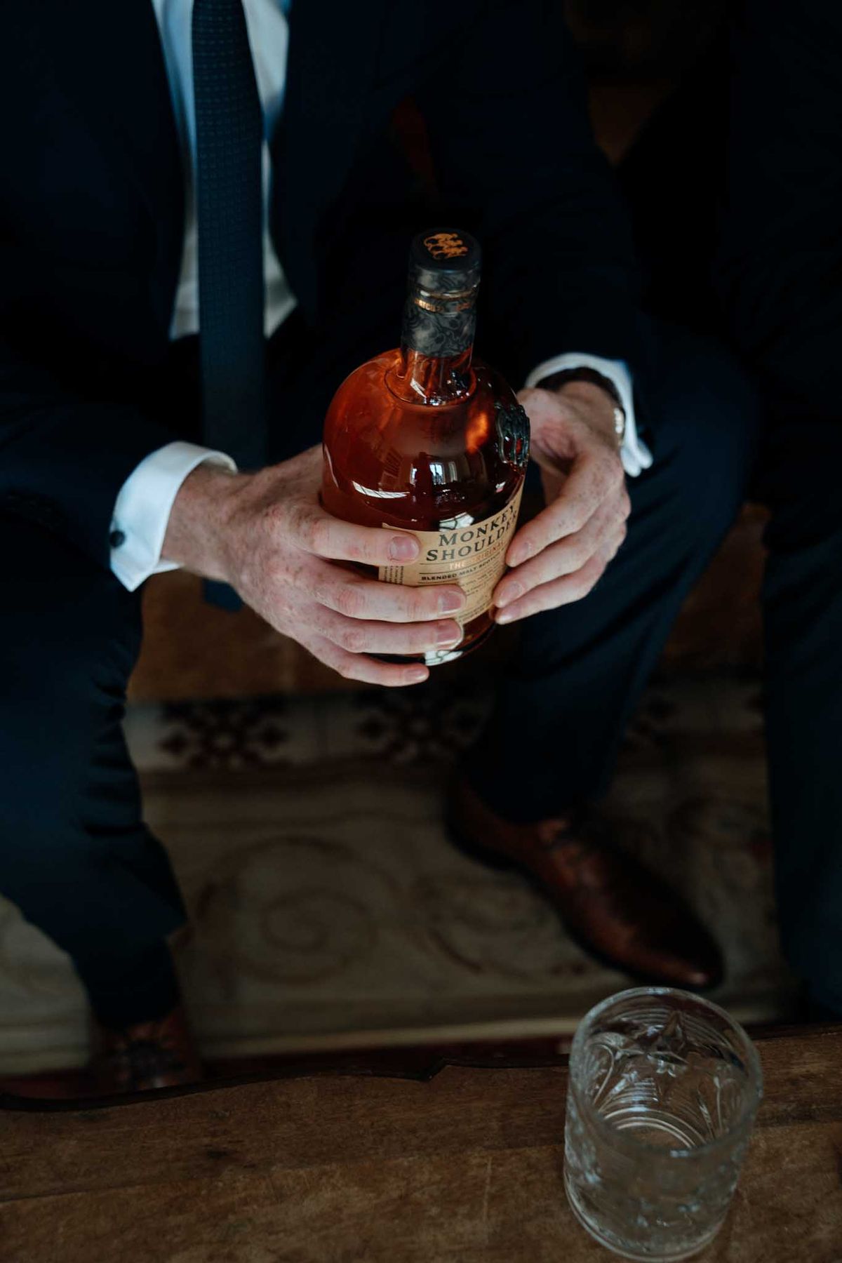 A close-up detail shot taken during the groom's getting-ready preparations, showing a man in a navy suit with a navy tie and white dress shirt holding a bottle of Monkey Shoulder blended malt Scotch whisky with both hands. A cut-crystal glass sits on a wooden surface in the foreground, and brown leather dress shoes are visible in the background beside another figure also in a dark suit. The image has a dark, moody tone with low ambient lighting and warm amber tones from the whisky bottle contrasting against the dark suits.
