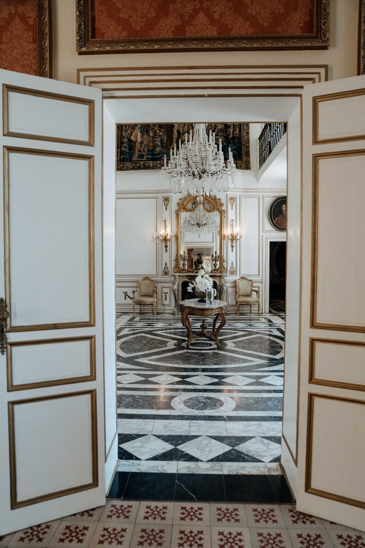 A wide shot looking through open double doors with white and gold-trim paneling into a grand chateau interior reception room. The room features a black and white geometric marble floor, white boiserie walls with gold molding details, a large crystal chandelier with candle-style arms, a gilt-framed overmantel mirror above a marble fireplace flanked by gold wall sconces, and two gold-framed Louis XVI-style upholstered chairs. A gilt carved occasional table in the center holds a vase of white flowers, likely white orchids or peonies. A large tapestry hangs above the doorway and a portrait painting is visible on the right wall. The foreground floor shows a red and cream patterned encaustic tile. No people are present in the image. Potential venue feature image.