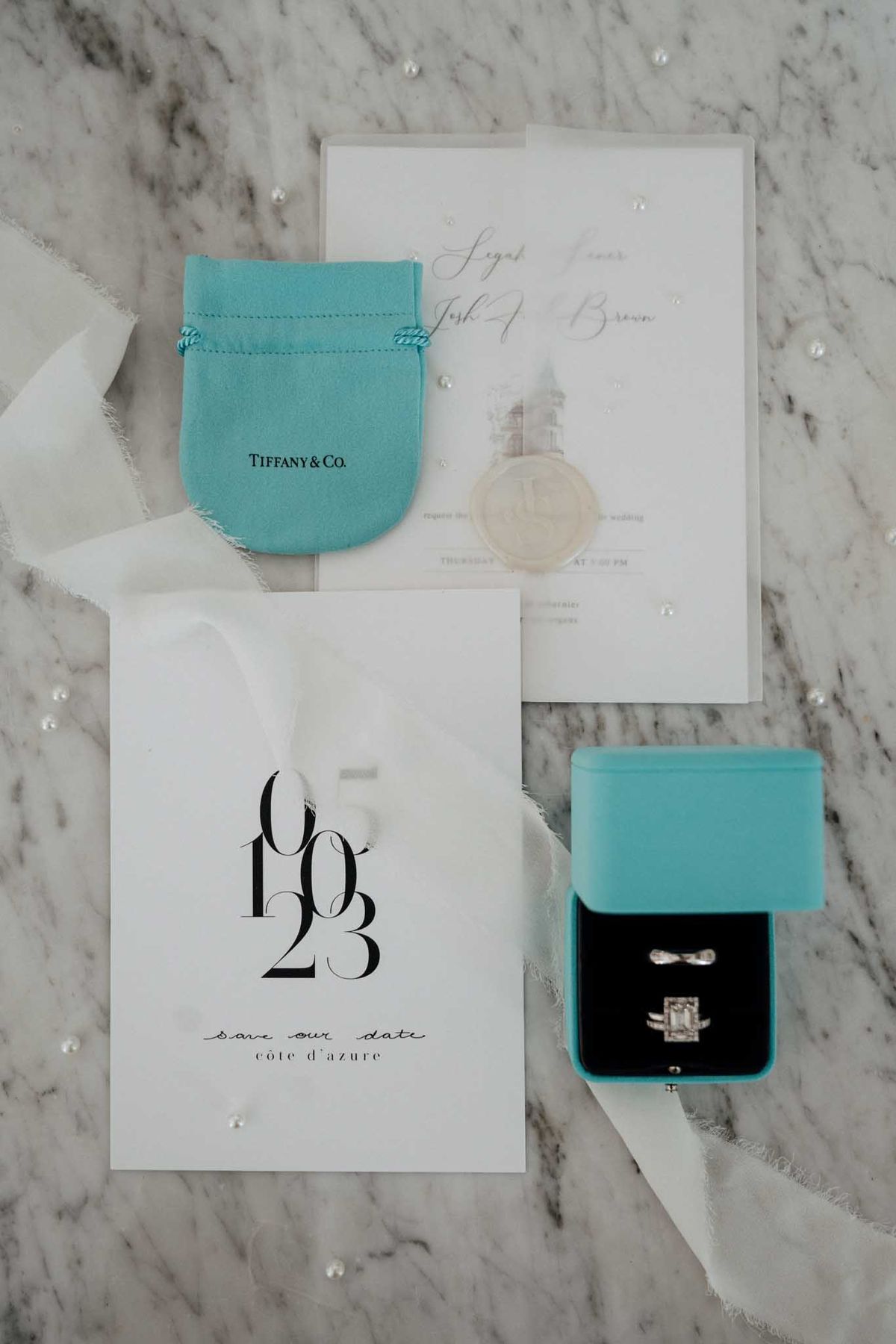 A flat lay detail shot of wedding stationery and jewelry accessories arranged on a white marble surface with scattered loose pearls and ivory silk ribbon. The stationery suite includes a white save-the-date card printed with the date 06/10/23 and the text 'save our date / côte d'azur' in black serif and script typography, alongside a white wedding invitation featuring gold calligraphy script and a château illustration, sealed with a cream wax stamp. A robin's-egg blue Tiffany & Co. velvet pouch sits alongside an open Tiffany blue ring box with a black velvet interior, displaying a diamond engagement ring with an emerald-cut center stone and a matching diamond wedding band, plus what appear to be two additional slim bands. The overall styling palette is Tiffany blue, white, and gold with a clean, modern-classic aesthetic.