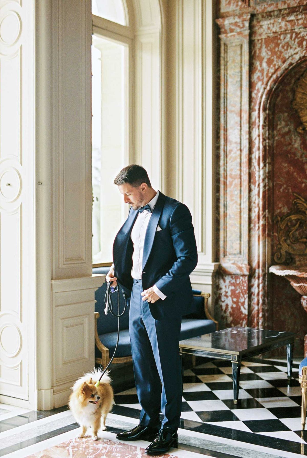 Groom in navy tuxedo with Pomeranian on leash in marble-floored chateau salon with gilded fireplace