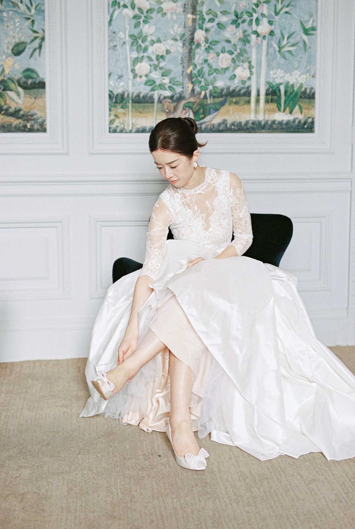 Bride seated putting on white bow-detail heels wearing lace-sleeve ball gown in chateau room with botanical panels
