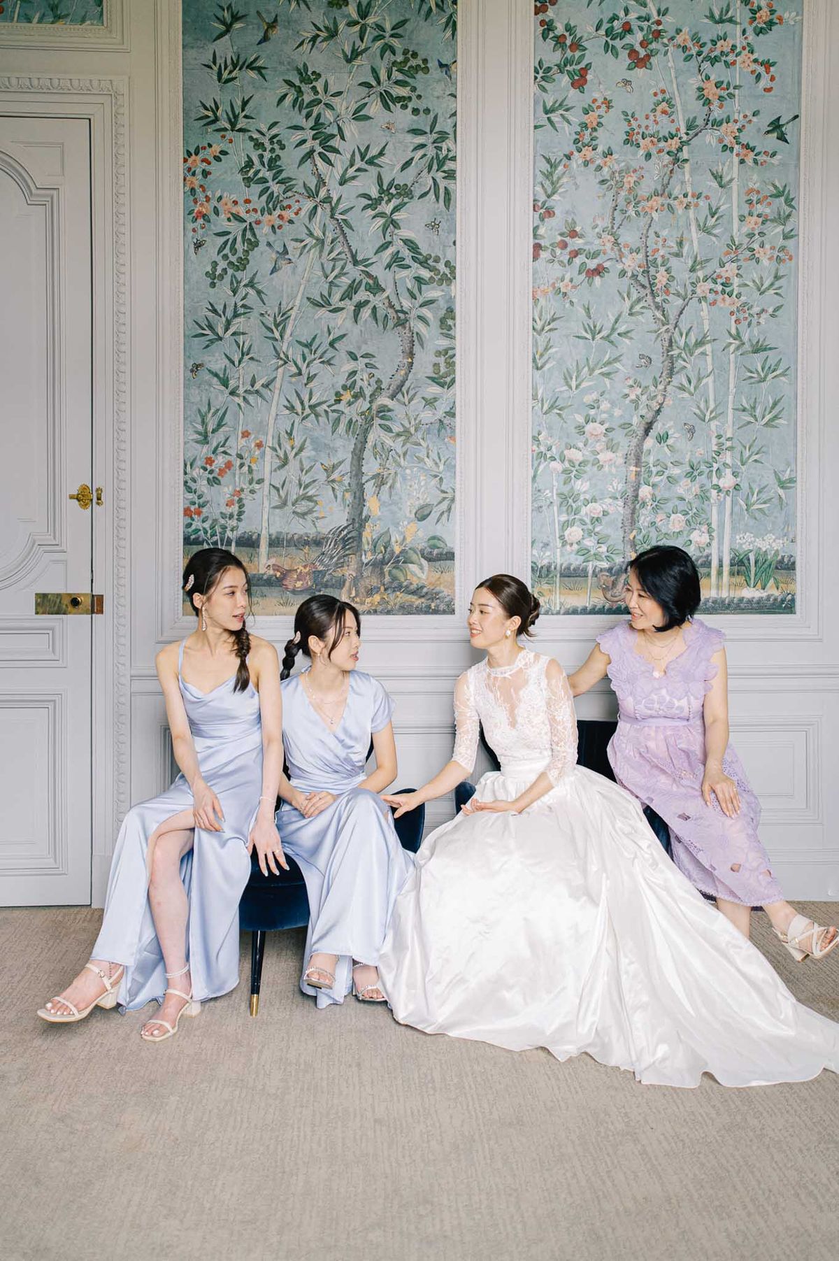 A bridal party portrait taken indoors, showing four women seated together on a navy velvet bench in a classically decorated room with white boiserie paneling and large chinoiserie-style mural panels depicting trees, botanicals, and birds in soft blue-green, coral, and cream tones. The bride sits third from the left wearing a full-ballgown skirt in white with a lace illusion high-neck long-sleeved bodice, her hair in an updo. Two bridesmaids wear periwinkle-blue satin dresses — one in a cowl-neck slip style and one in a short-sleeve wrap style — while a fourth woman, likely the mother of the bride, wears a short lilac lace dress with ruffle cap sleeves. The women are engaged in relaxed conversation, smiling at one another. The overall palette of the group — white, periwinkle, and lilac — coordinates with the cool blue tones of the mural backdrop. This is a wide portrait shot with a classic, polished indoor styling approach.