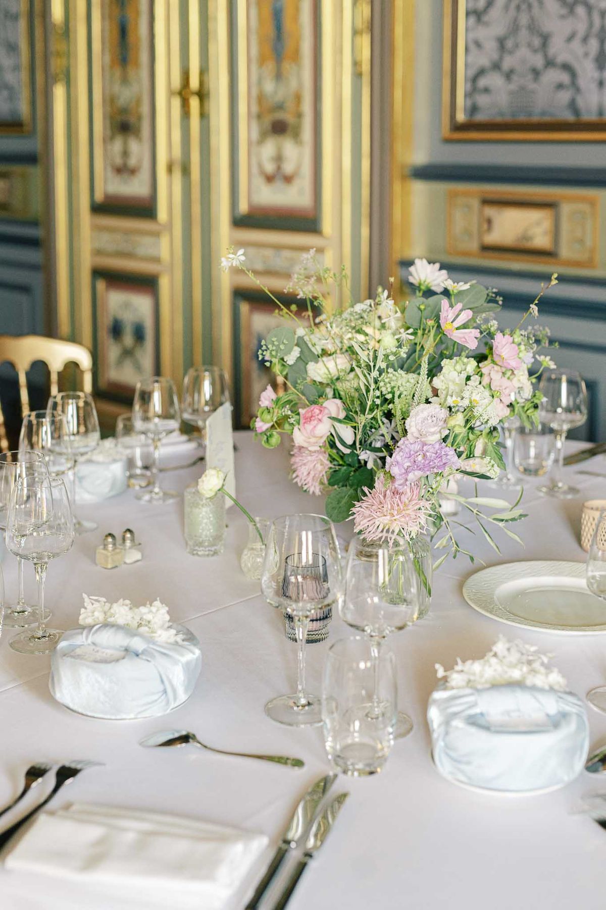 Wedding reception table with crystal glasses, sage charger plates, pink dahlia and lavender peony centerpiece in a gilded ...